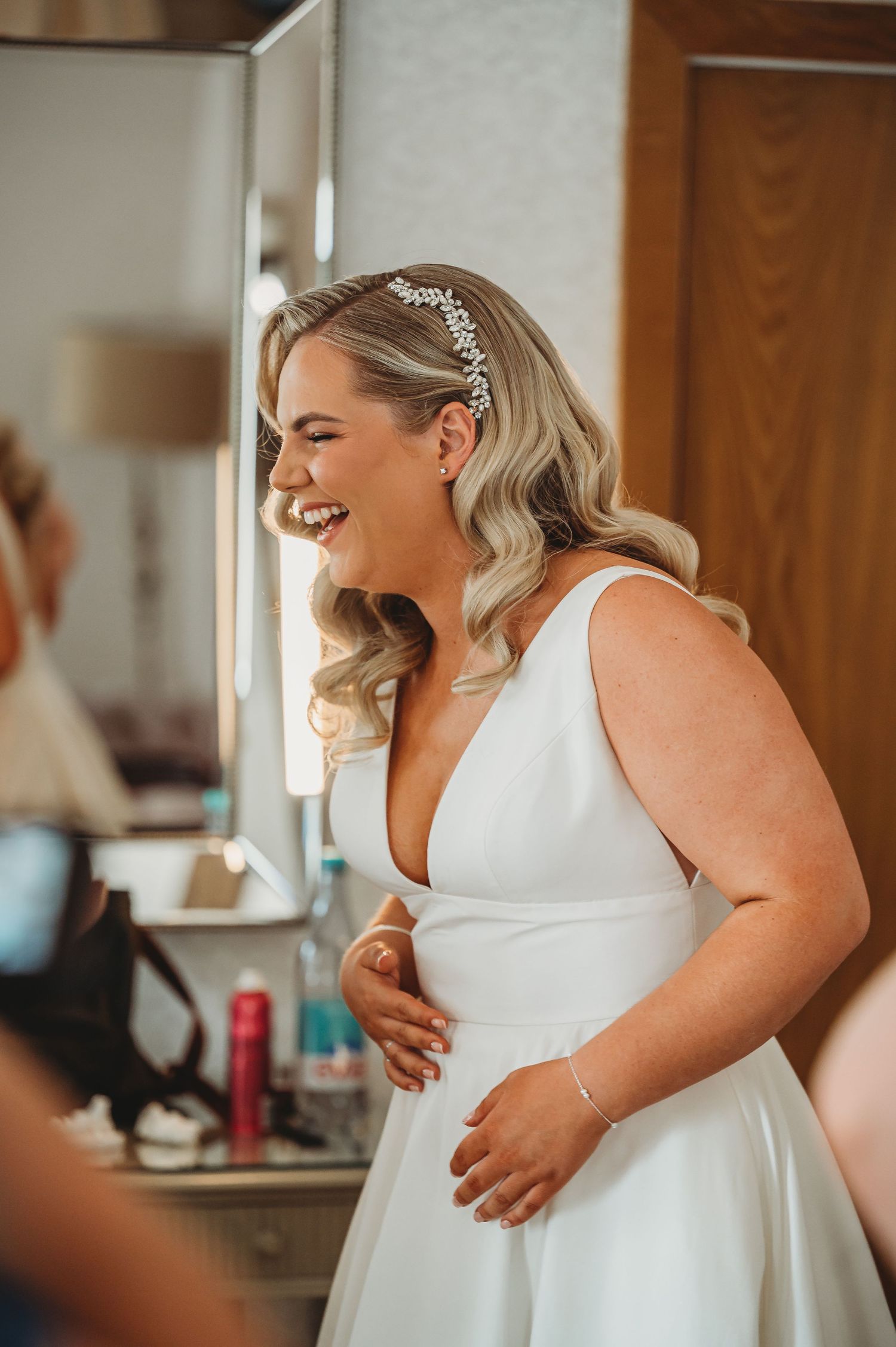 A bride wearing a classic white wedding dress and crystal headpiece laughs joyfully while getting ready for her ceremony.