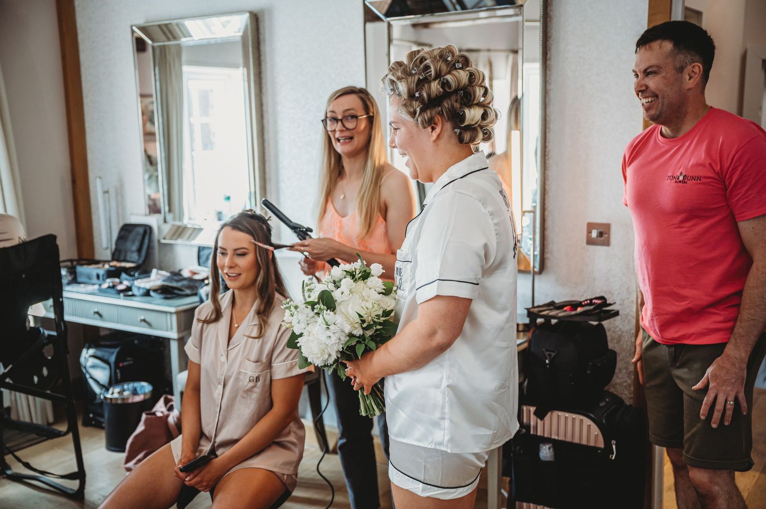 A candid moment in a bright salon shows people sharing laughter while getting ready with flowers and styling equipment.