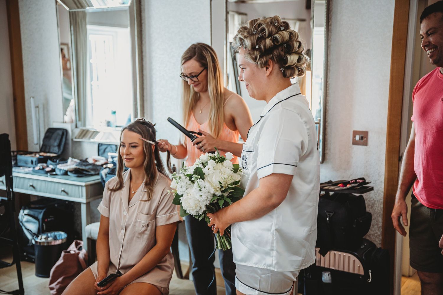 Hair stylist and team prepare for wedding with floral bouquet in bright getting ready room.