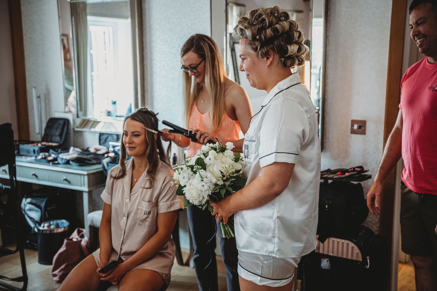 Wedding preparation scene in a home with stylists doing hair while holding a white floral bouquet.
