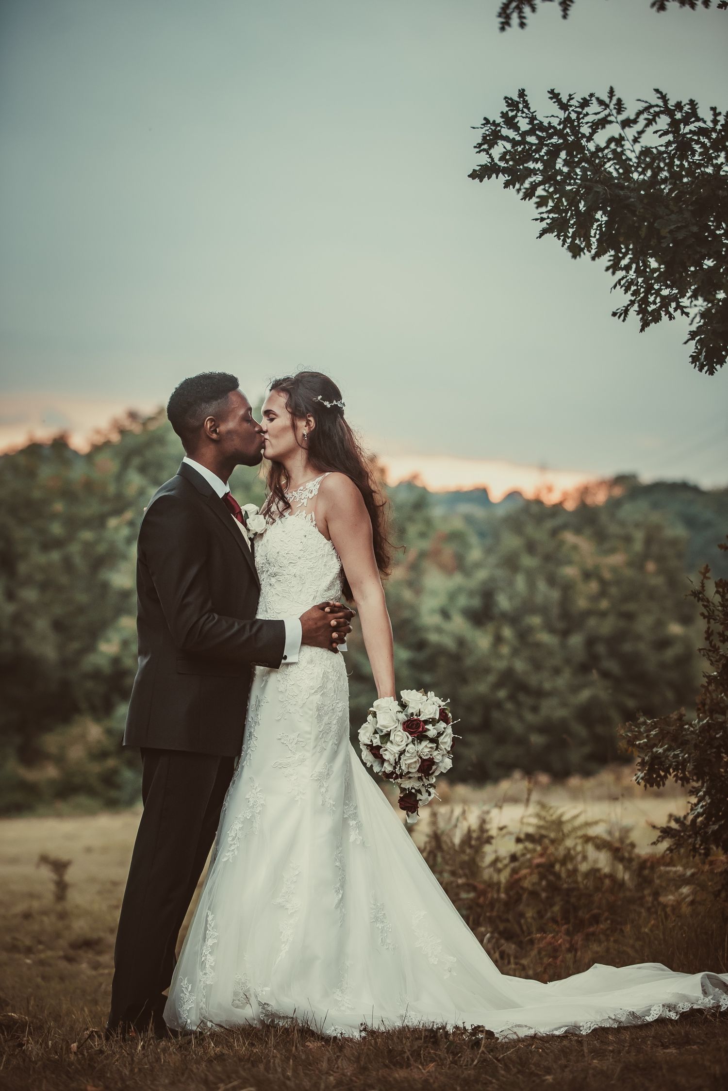 A romantic wedding couple embraces outdoors at sunset with rolling hills and trees in the background.