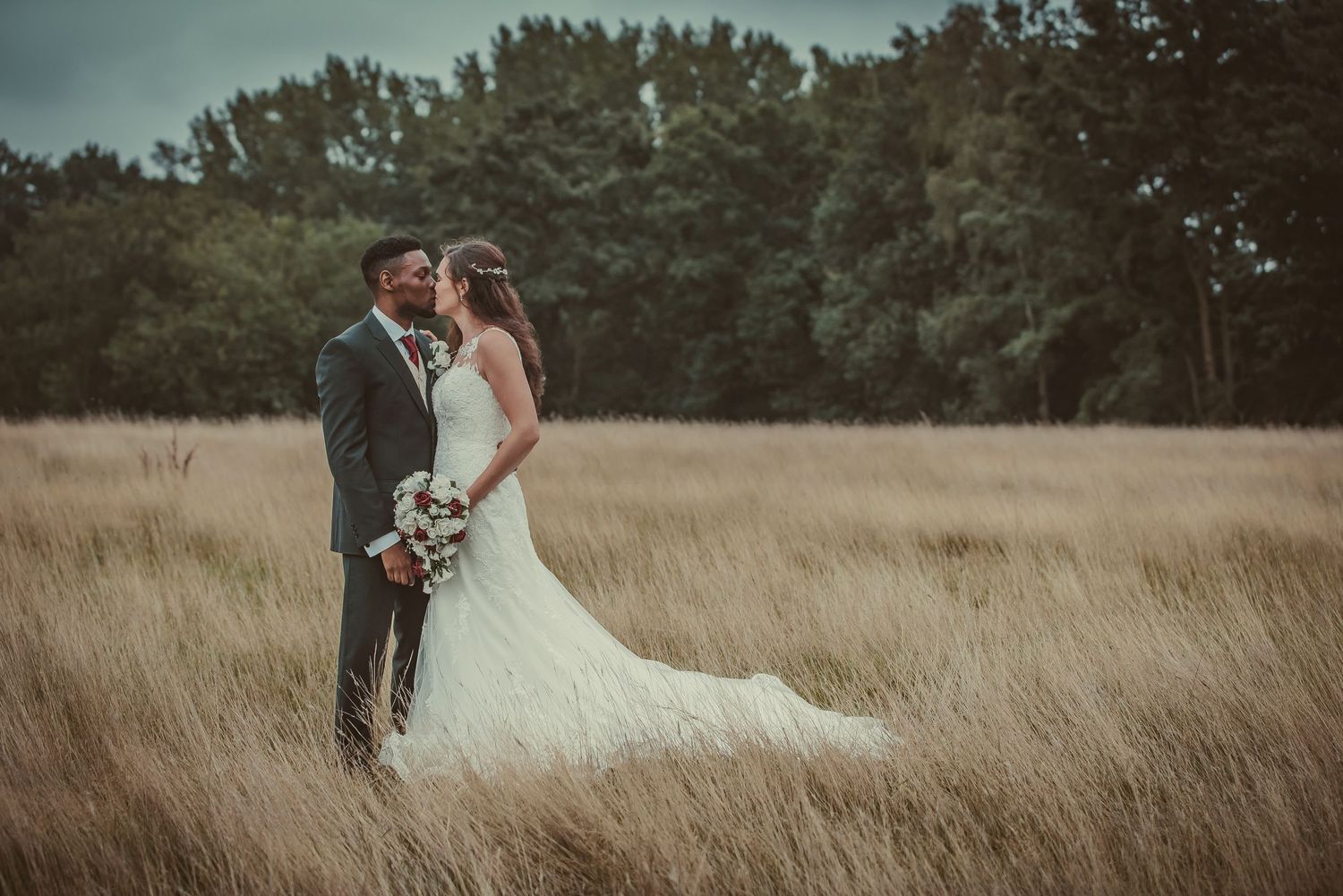 A newlywed couple stands close together in a golden wheat field with evergreen trees in the background.