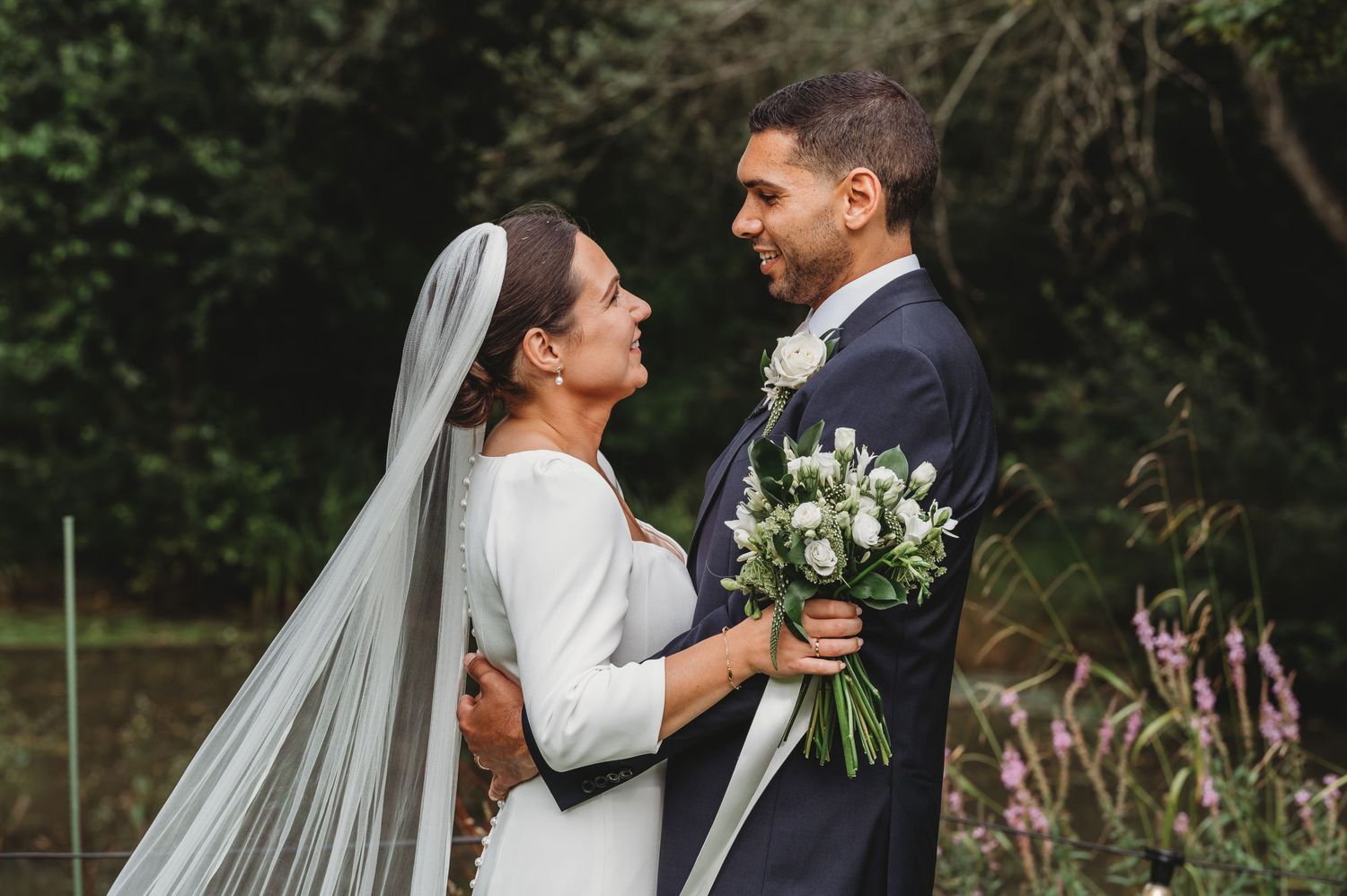 A joyful couple in wedding attire share a candid moment while holding a white floral bouquet outdoors.