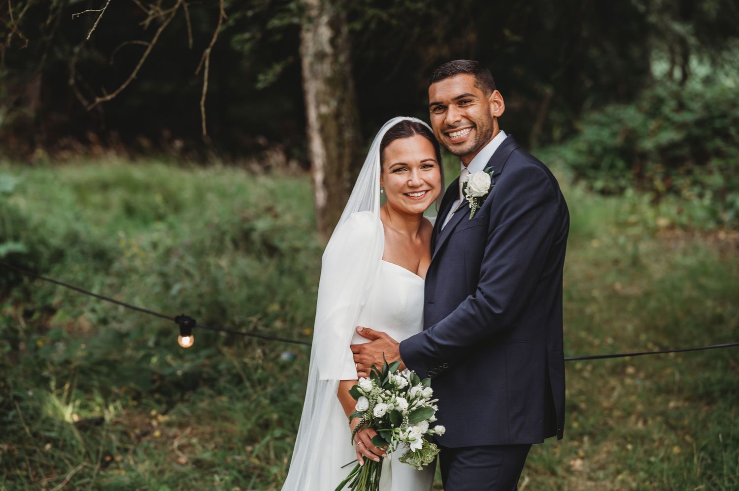 A newlywed couple embraces outdoors in a wooded garden setting with the bride holding a white floral bouquet.