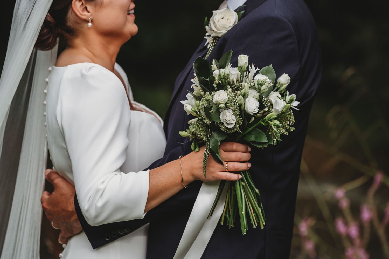 A wedding couple embraces while holding an elegant white rose and greenery bouquet at their wedding celebration.