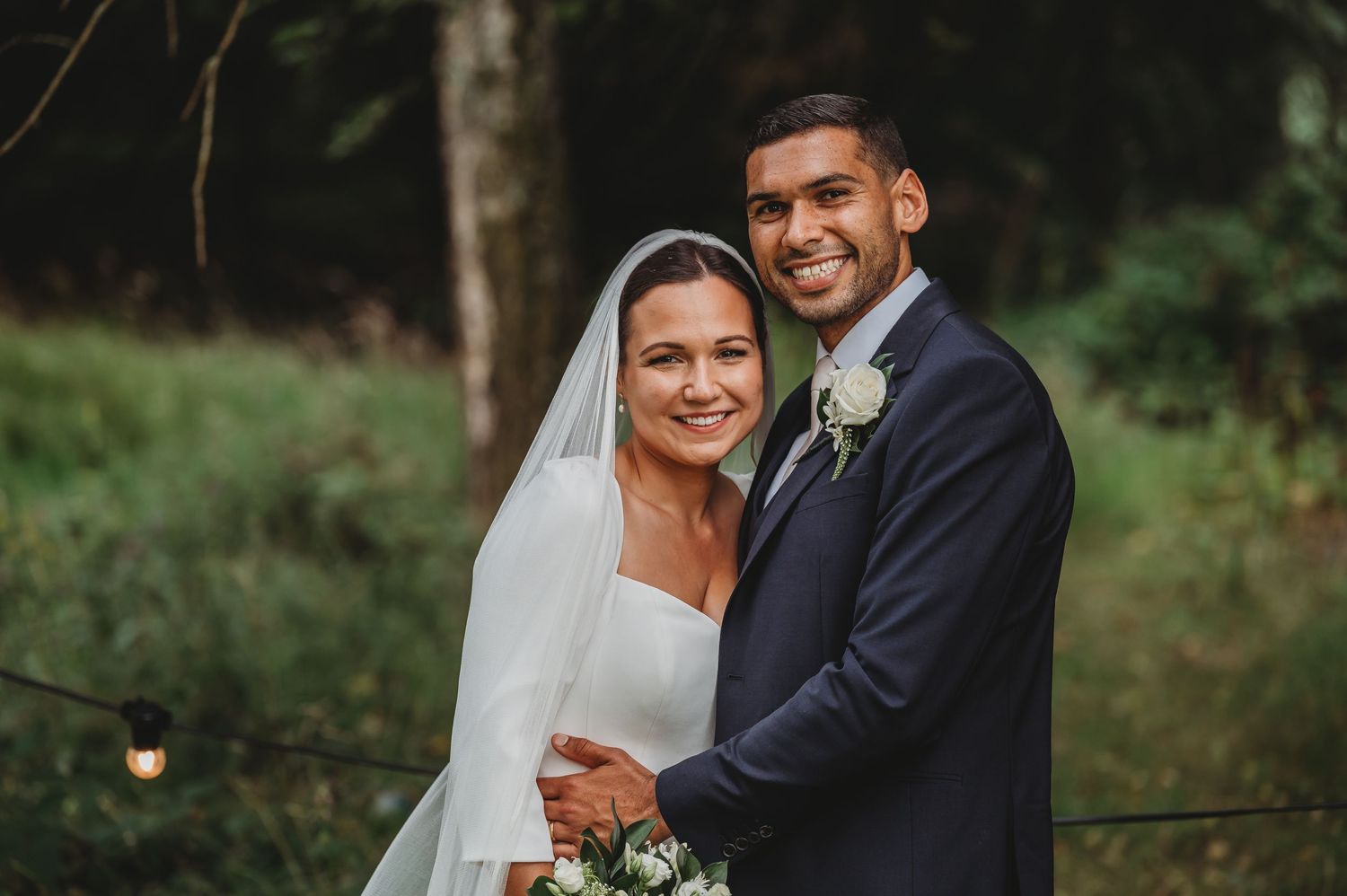A happy couple poses together outdoors during their wedding celebration in a natural wooded setting.
