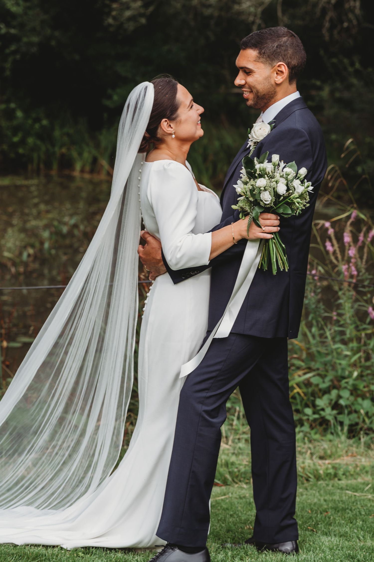 A couple wearing a white wedding dress and navy suit embrace while holding a bouquet of white flowers outdoors.
