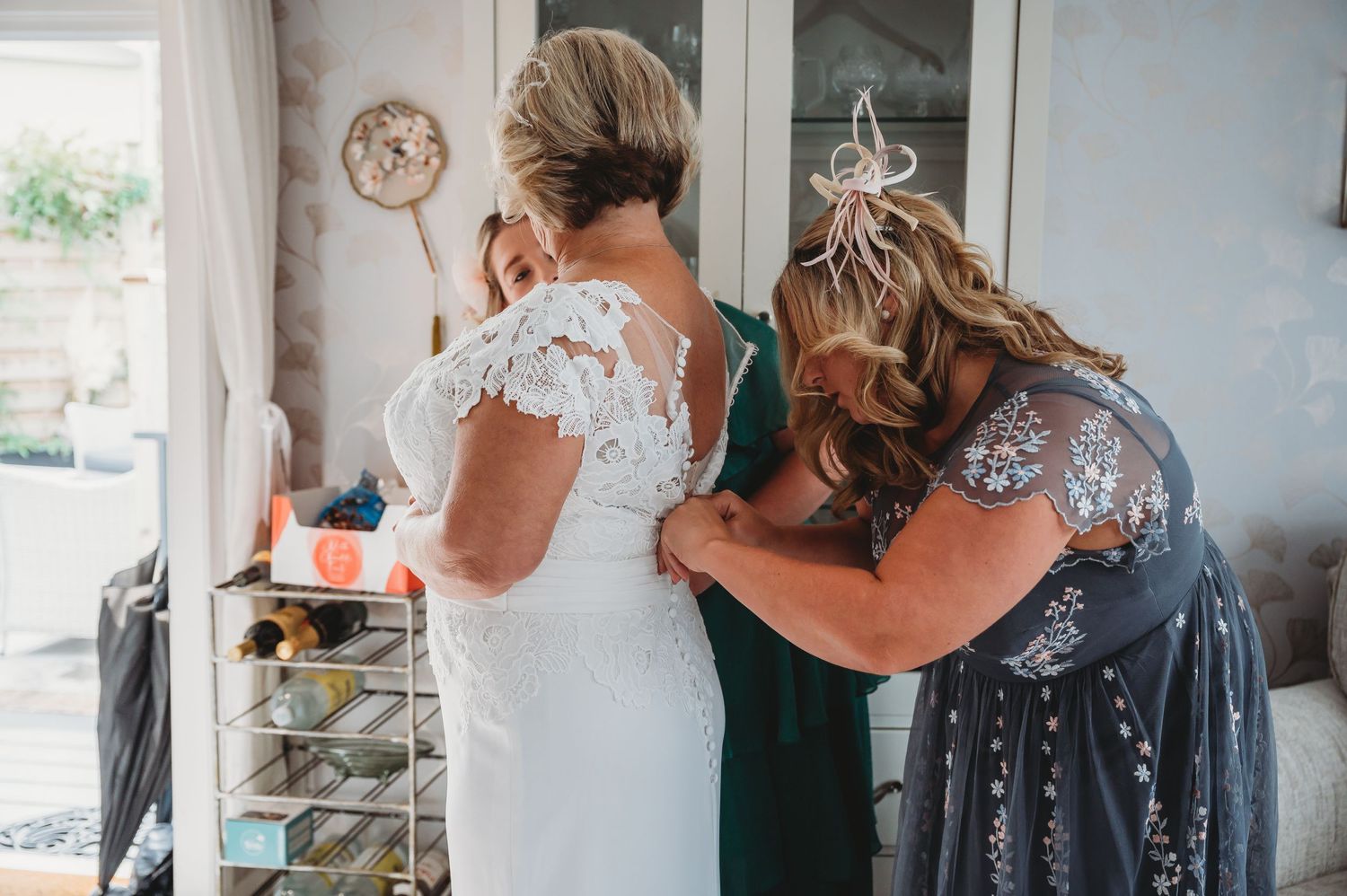 Two people in formal attire assist with adjustments to a white lace wedding dress in an indoor setting.