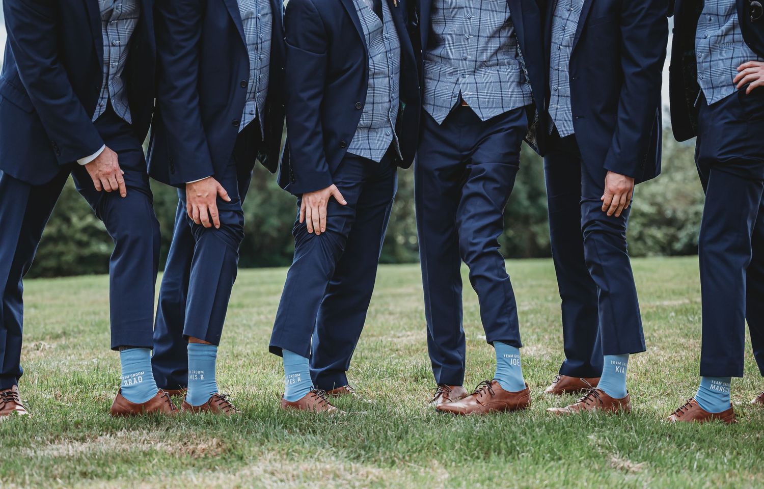 A group wearing matching dark suits and brown shoes stand together on grass showing just their legs and feet.
