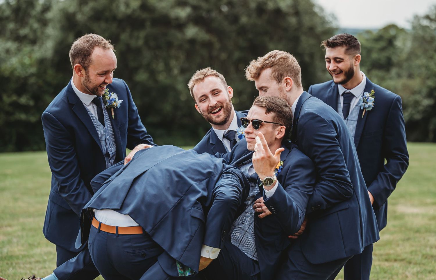 Groomsmen in navy blue suits sharing a candid laughing moment together on grassy lawn during wedding celebration.