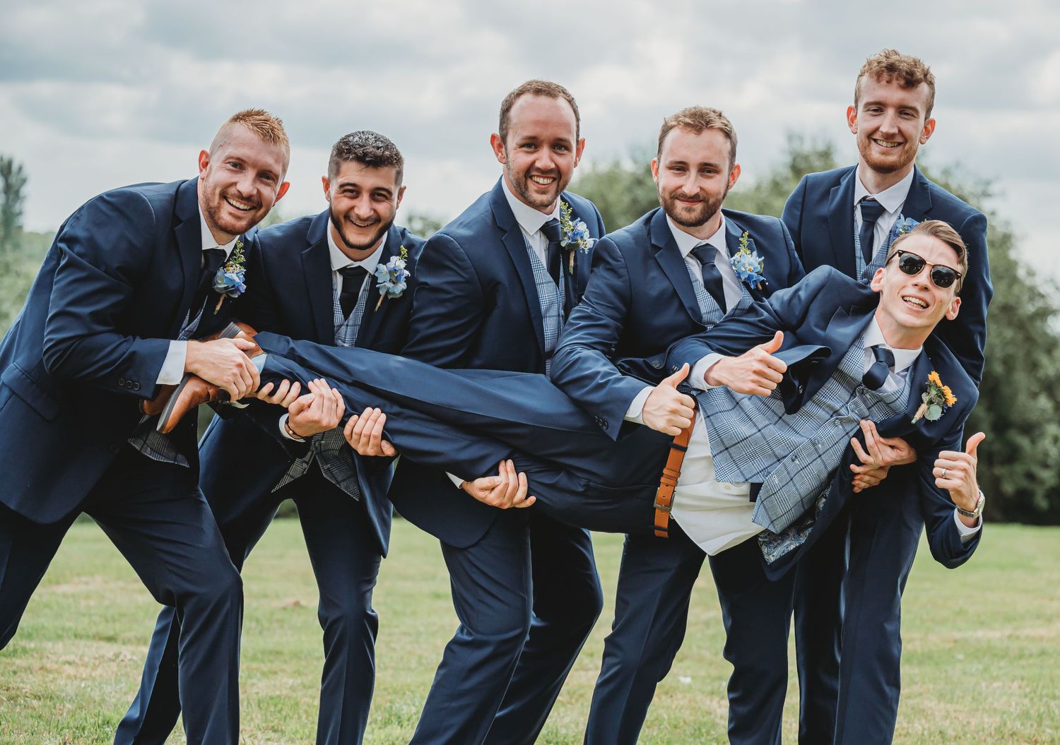 Groomsmen in navy suits pose playfully lifting a member of the wedding party on a grassy field outdoors.