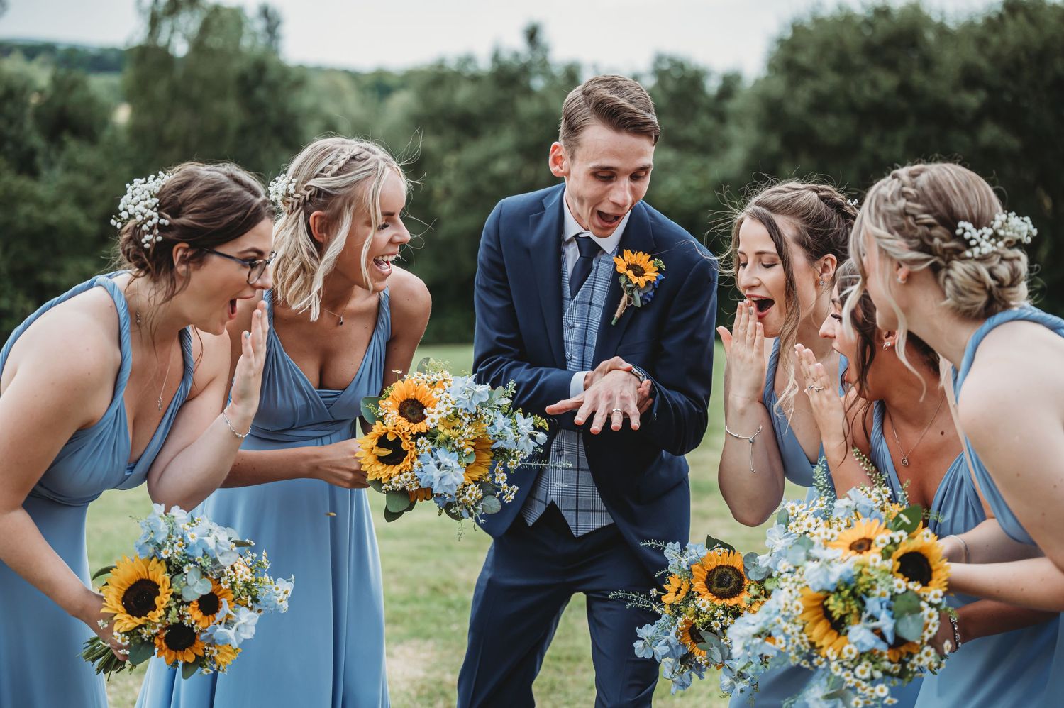 Wedding party in light blue dresses and navy suit share a lively moment with bright sunflower bouquets outdoors.