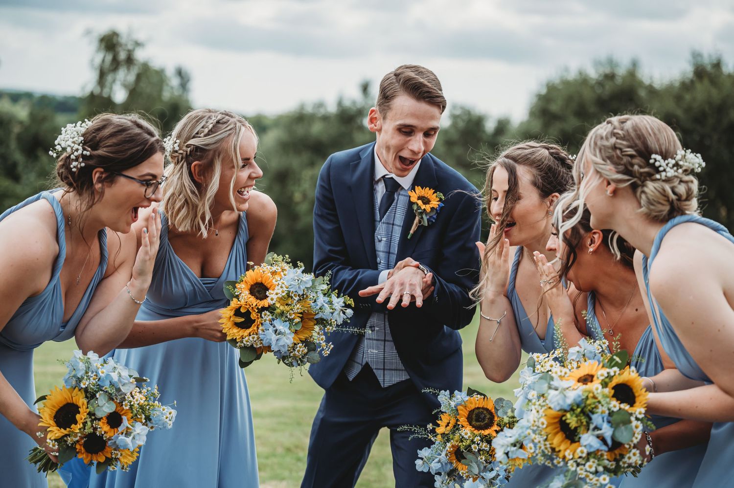 A wedding party in light blue dresses holding sunflower bouquets sharing a candid moment outdoors.