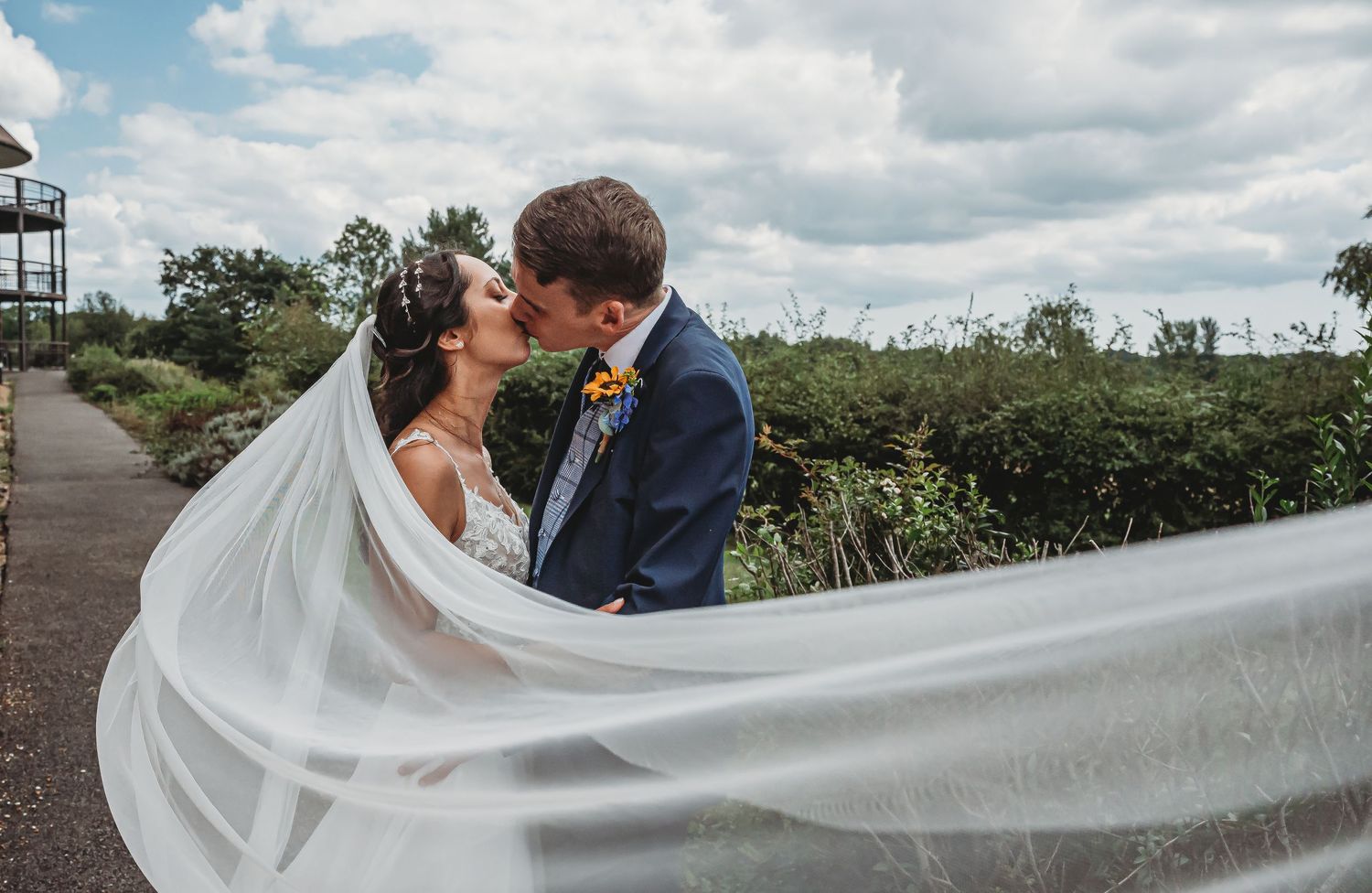 A bride and groom share a romantic kiss outdoors on their wedding day as her long white veil flows gracefully in the breeze.