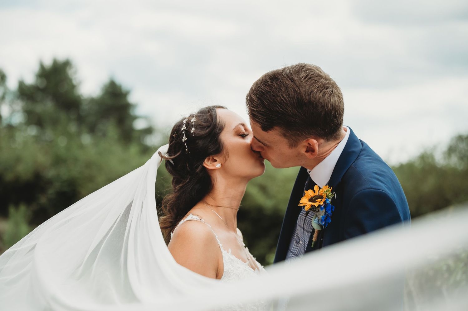 A romantic wedding couple shares a tender kiss outdoors as the bride's white veil flows in the breeze.