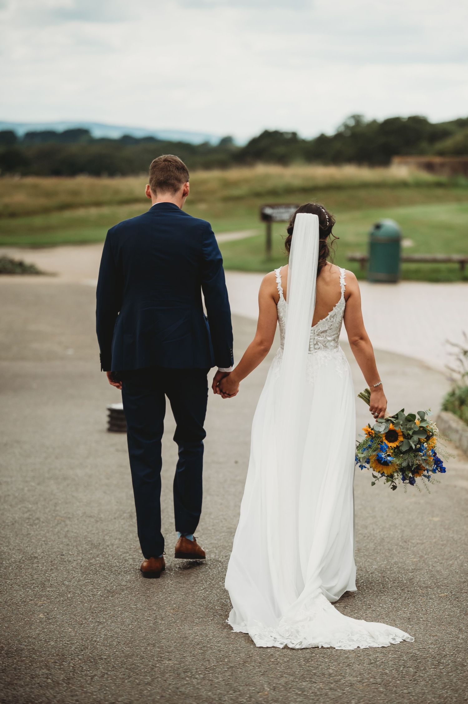 A newlywed couple walking hand in hand on a path while the bride holds a vibrant blue bouquet.