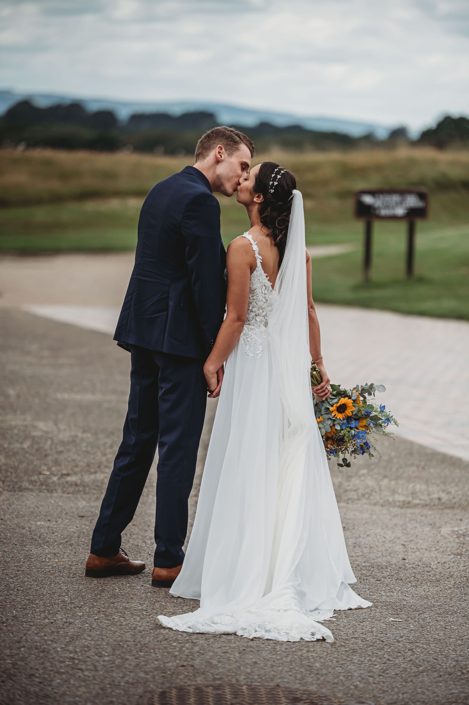A romantic wedding couple shares a tender moment on a path with mountains in the background and a colorful bouquet.