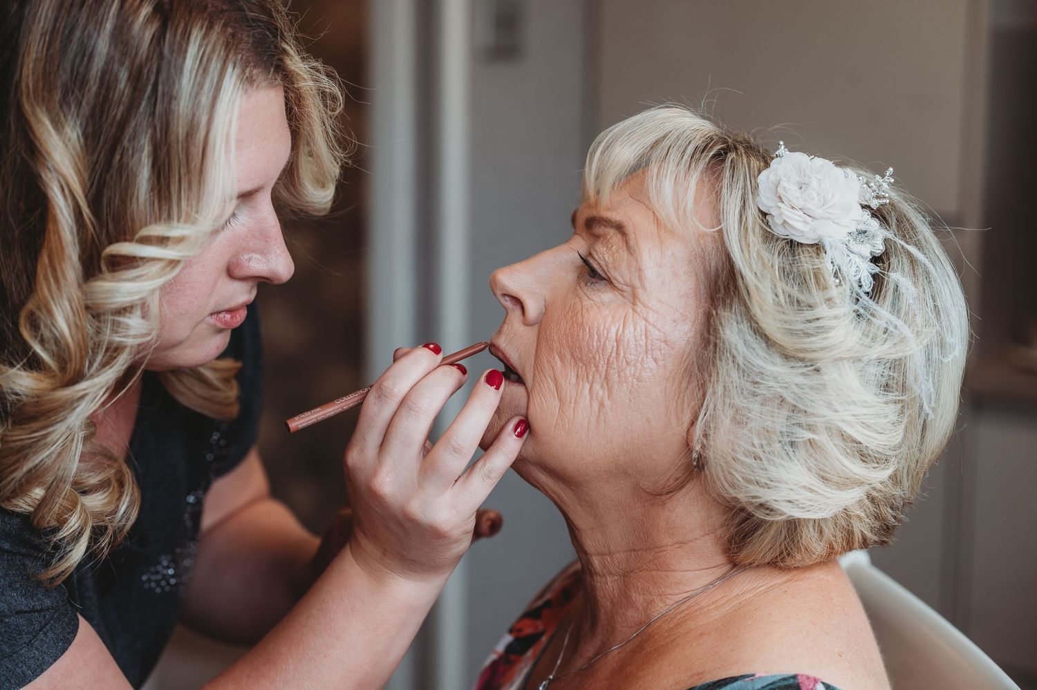 A makeup artist applies lipstick to a bride with a white floral hair accessory during wedding preparations.