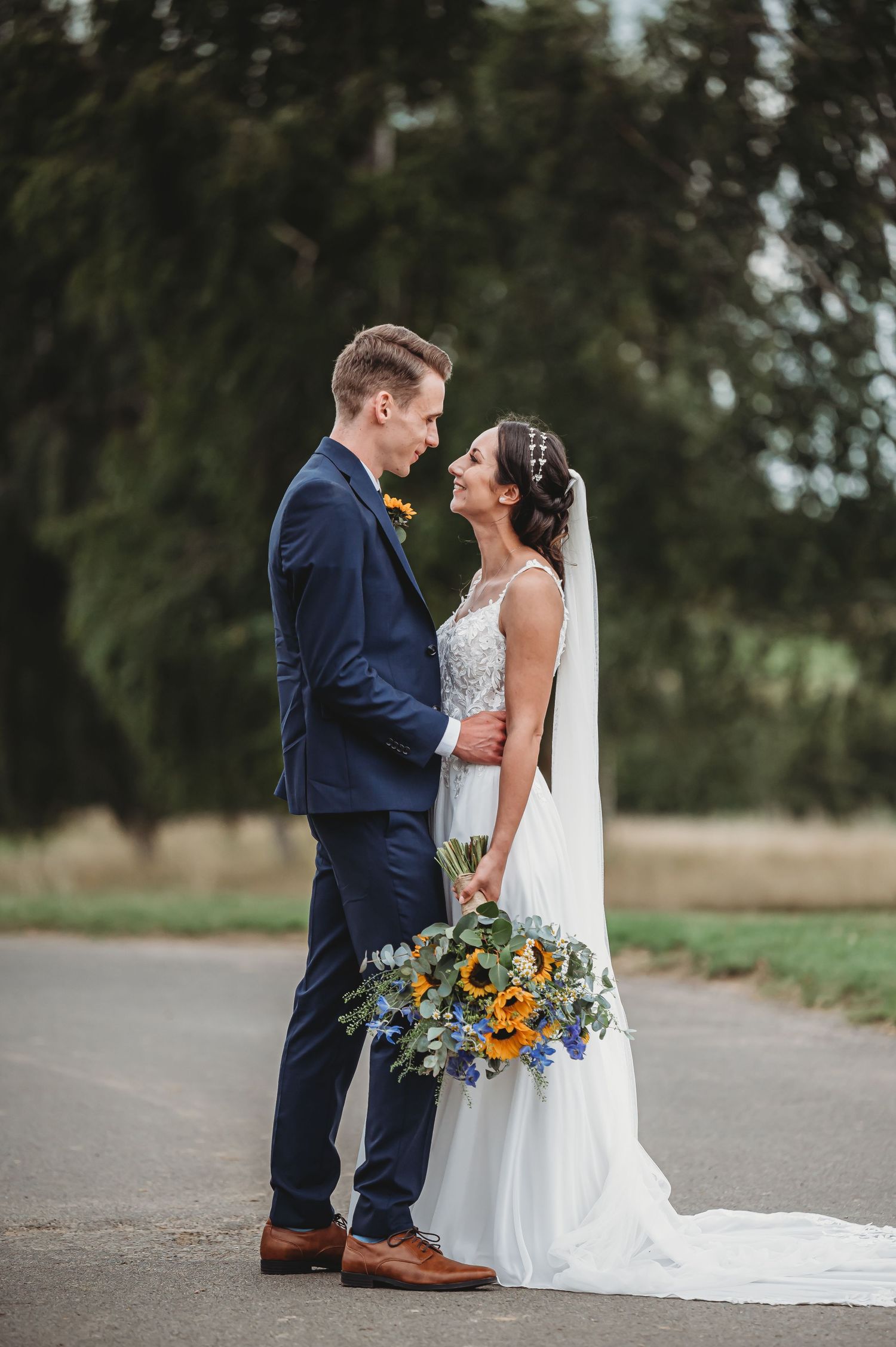 A couple in wedding attire share an intimate moment outdoors while holding a vibrant sunflower bouquet.