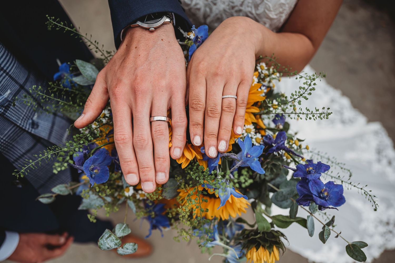 Two hands displaying wedding rings while holding a vibrant bouquet with sunflowers, blue flowers and greenery.