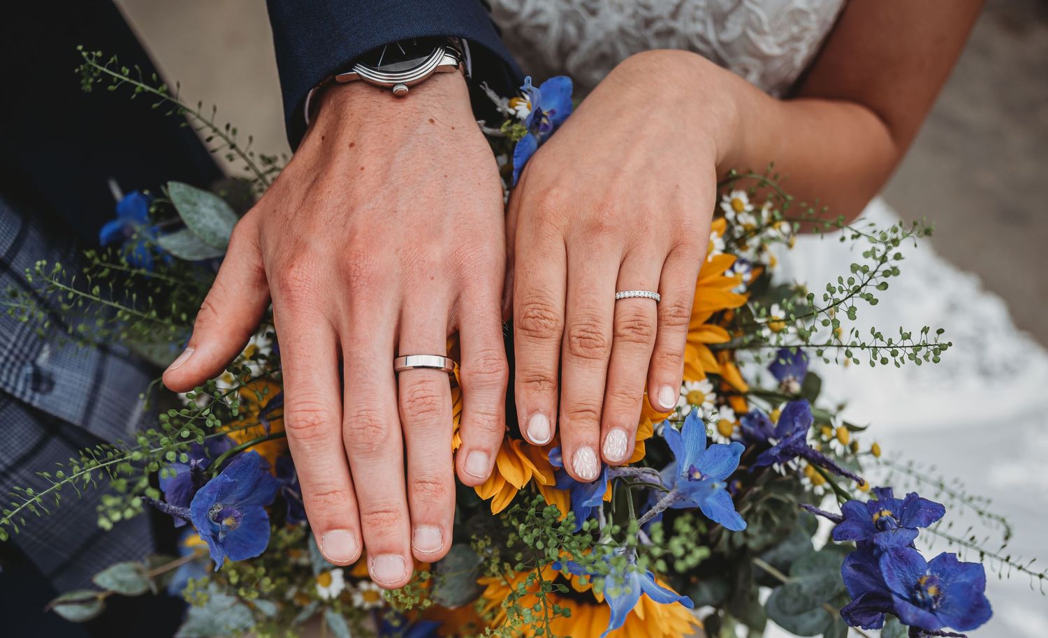 Two hands with wedding rings displayed above a colorful bouquet with blue and yellow flowers.