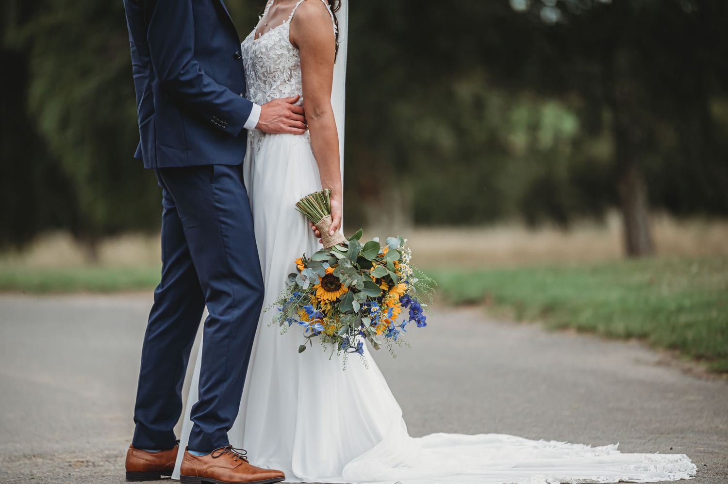 Wedding couple stands together outdoors while holding beautiful sunflower and wildflower bouquet.