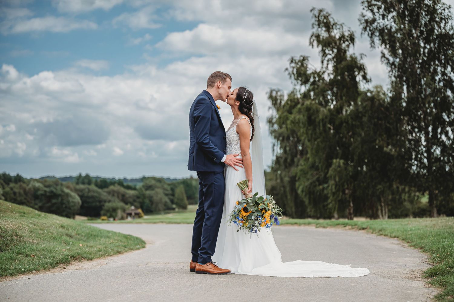 Two people share a romantic moment on a winding countryside path under a blue cloudy sky with green trees in background.