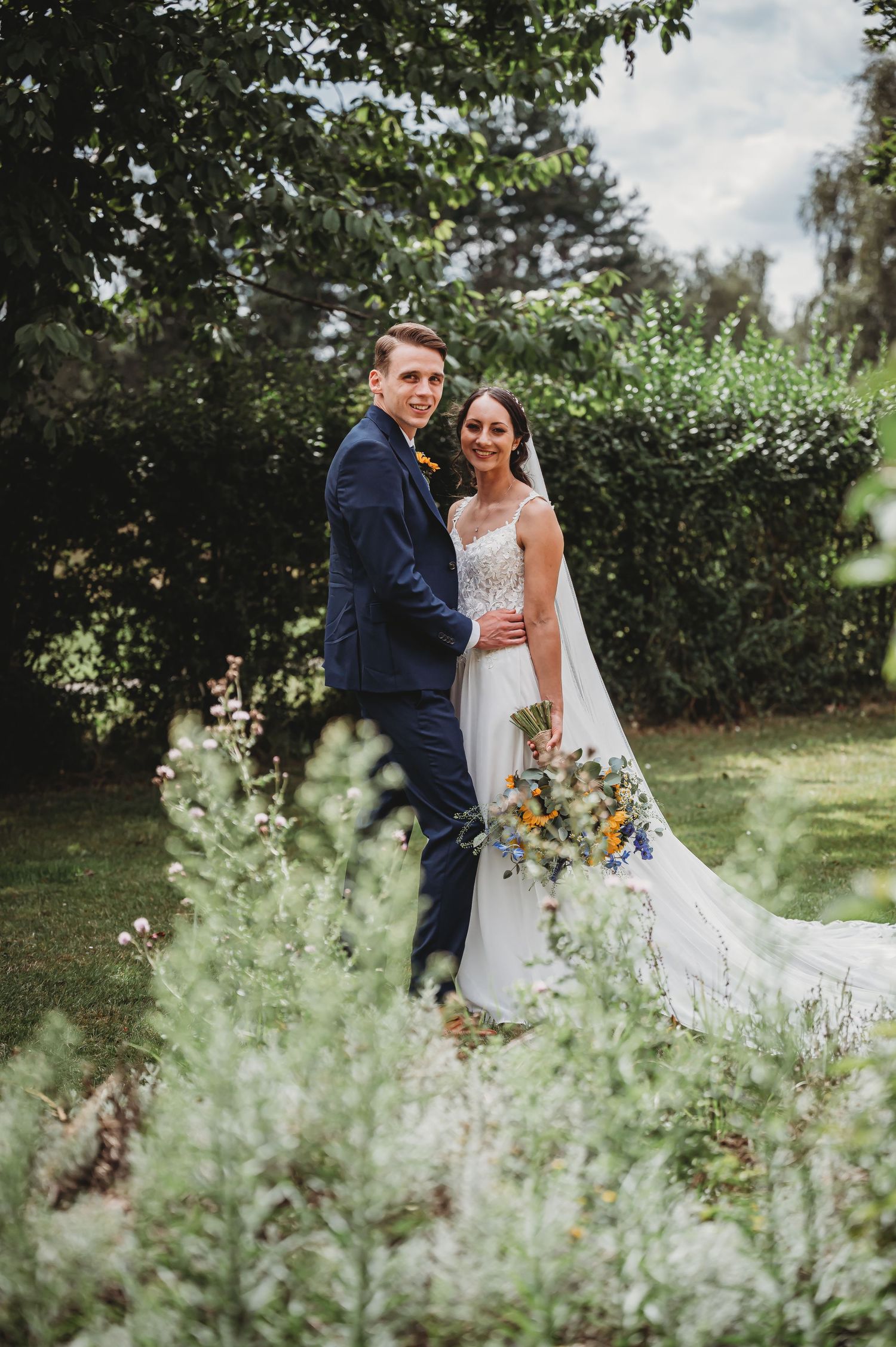 A wedding couple poses together in a garden setting surrounded by lush greenery and holding a yellow floral bouquet.