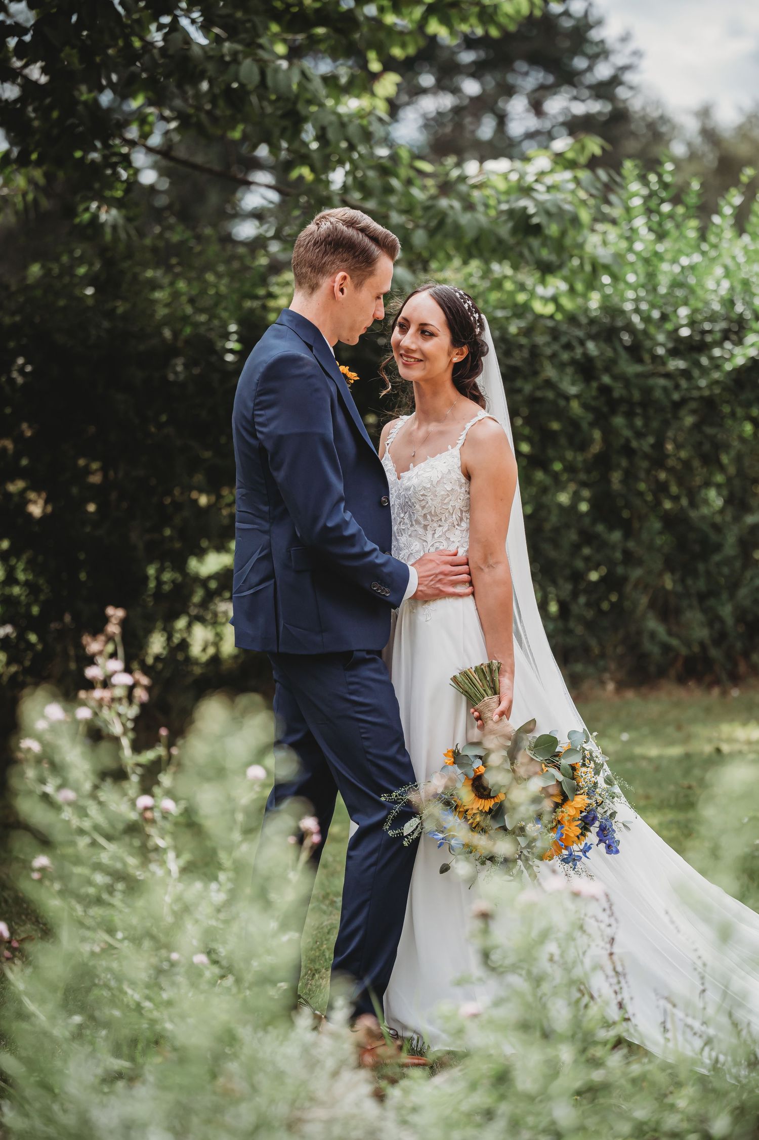 A bride in white lace gown holds a sunflower bouquet while standing with her groom in a navy suit in a garden setting.