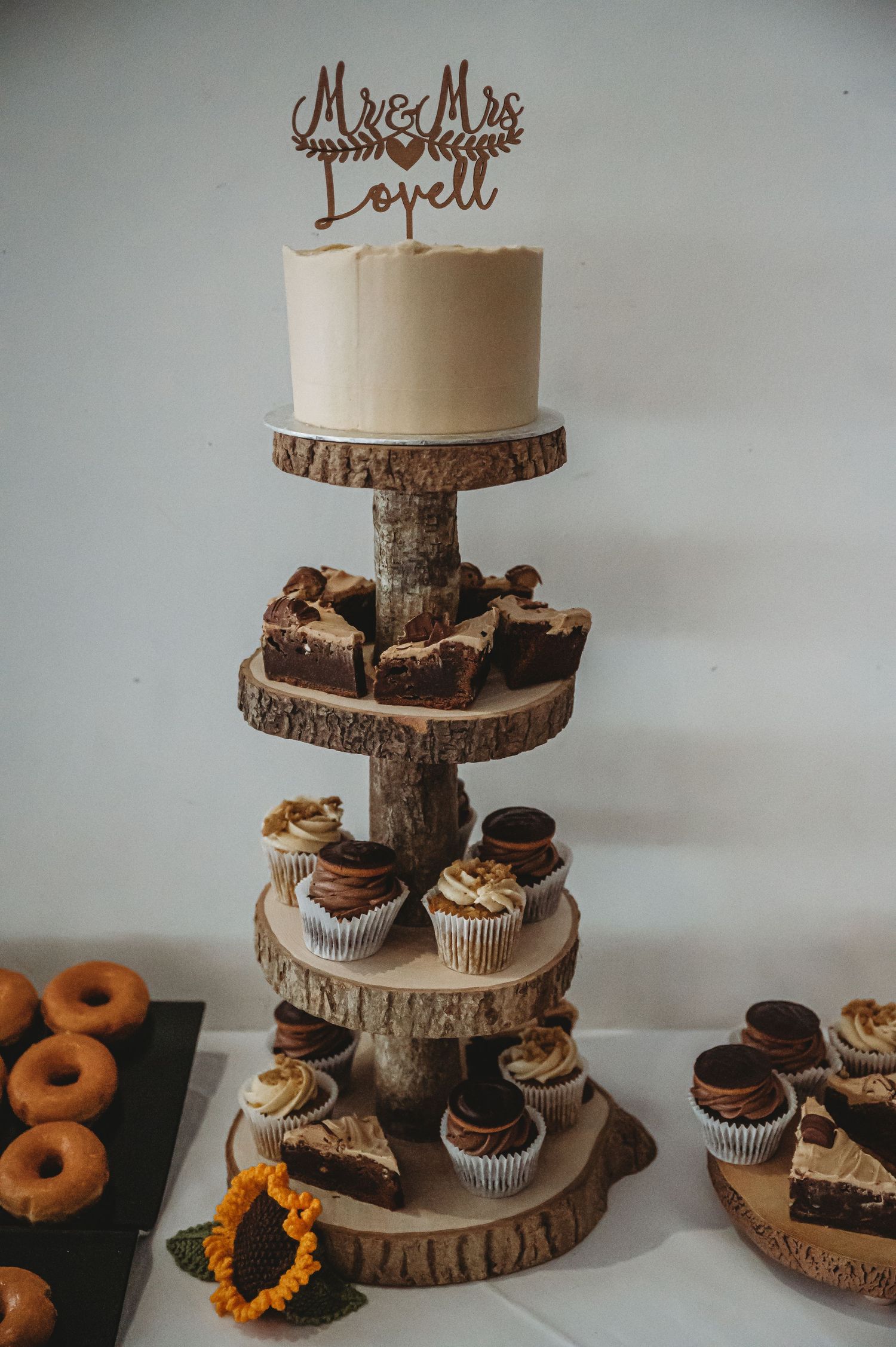 Rustic wood slice tiered cake stand displays white wedding cake and chocolate cupcakes with brown cake topper decoration.