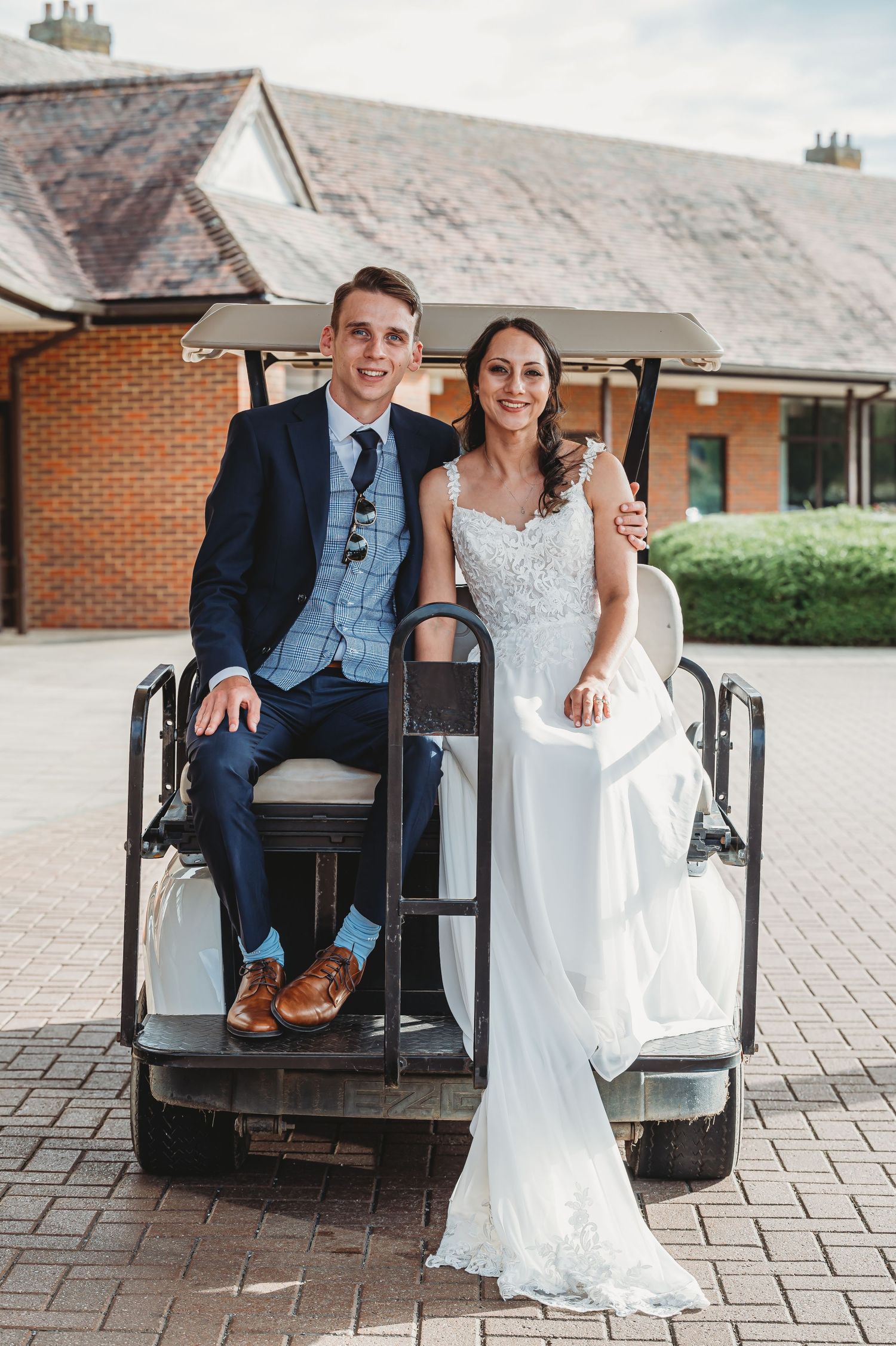 A couple in wedding attire sits on a golf cart outside a brick building on their special day.