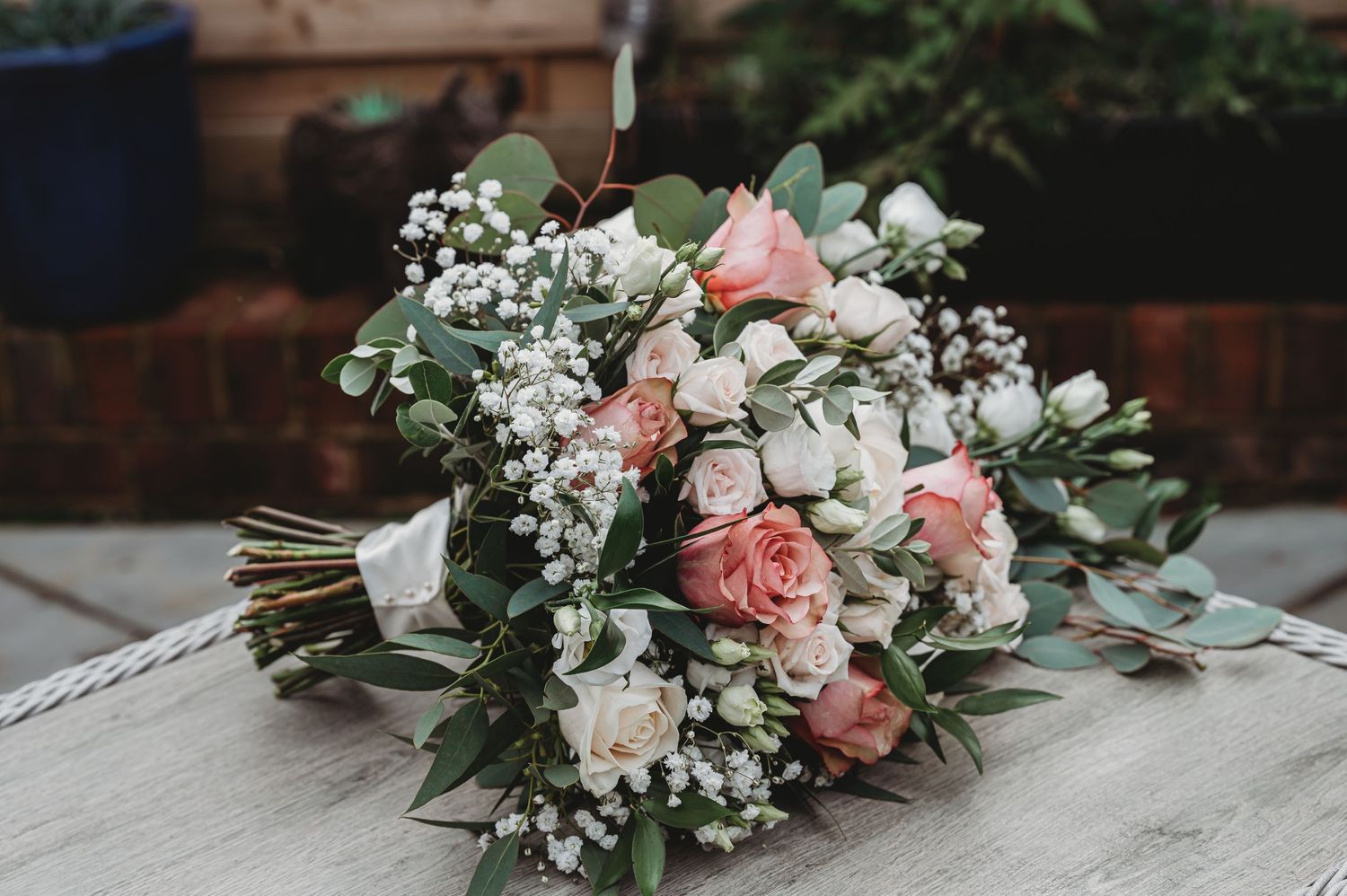 A romantic wedding bouquet featuring pink roses, white baby's breath, and eucalyptus leaves rests on a gray surface.