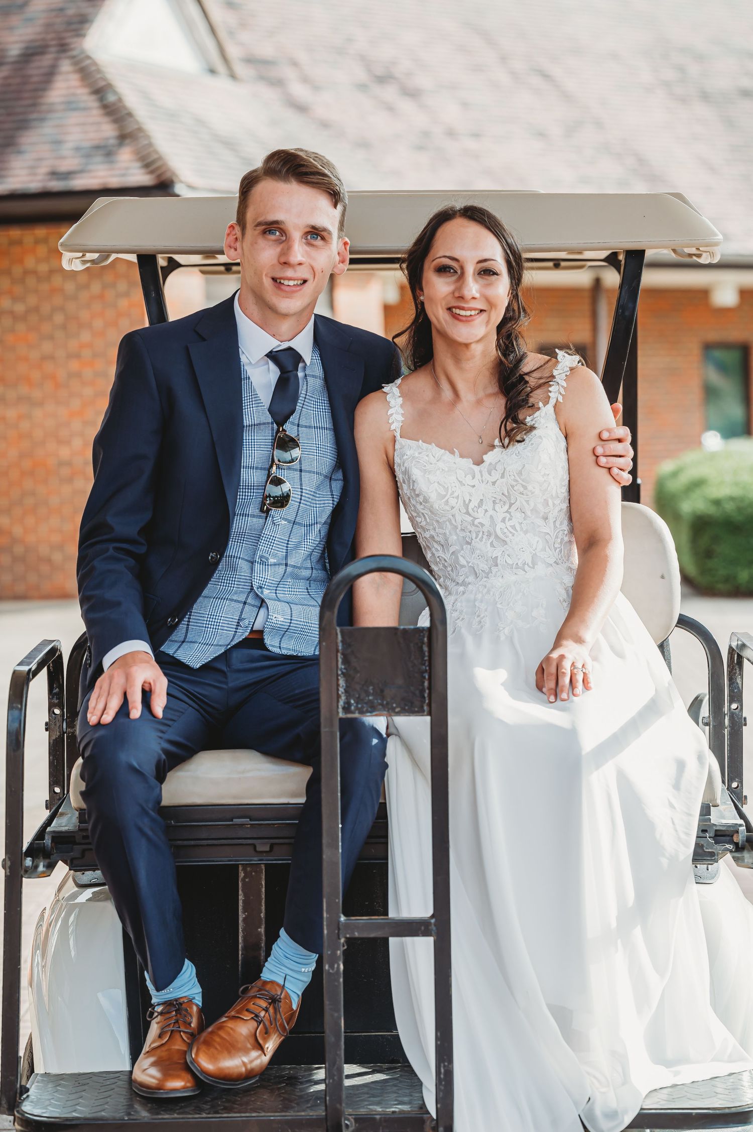 A couple sits together in a white golf cart on their wedding day, wearing formal blue suit and white bridal gown.