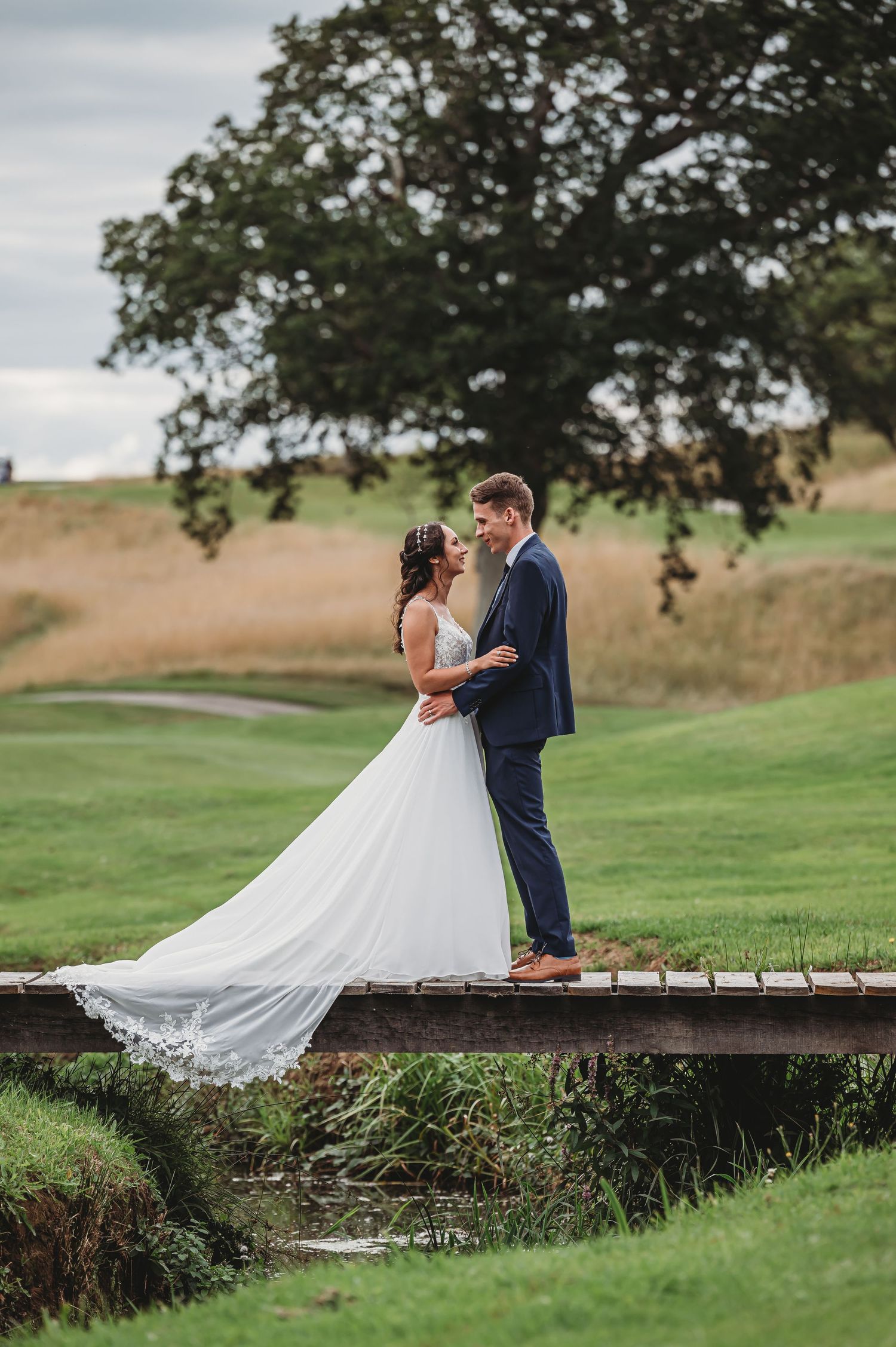 A bride and groom share an intimate moment on a rustic wooden bridge overlooking a scenic golf course landscape.