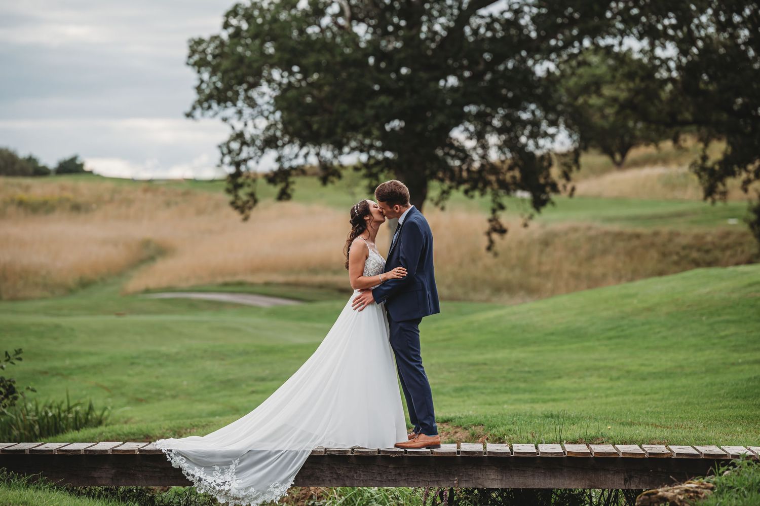 A couple in wedding attire embraces on a wooden bridge overlooking a scenic golf course with rolling hills in the background.