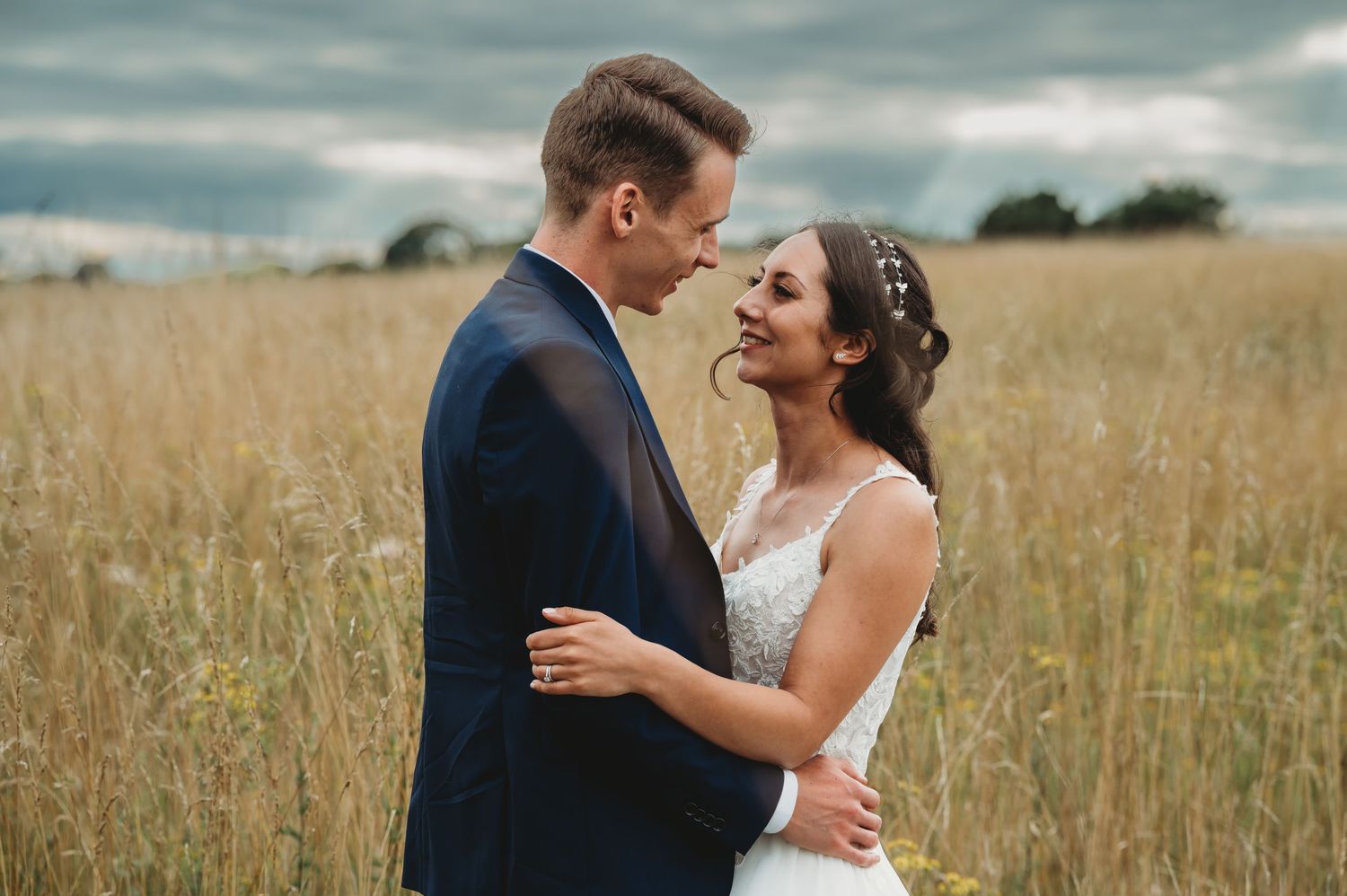 A bride and groom sharing an intimate moment in a sun-lit golden wheat field under cloudy skies.