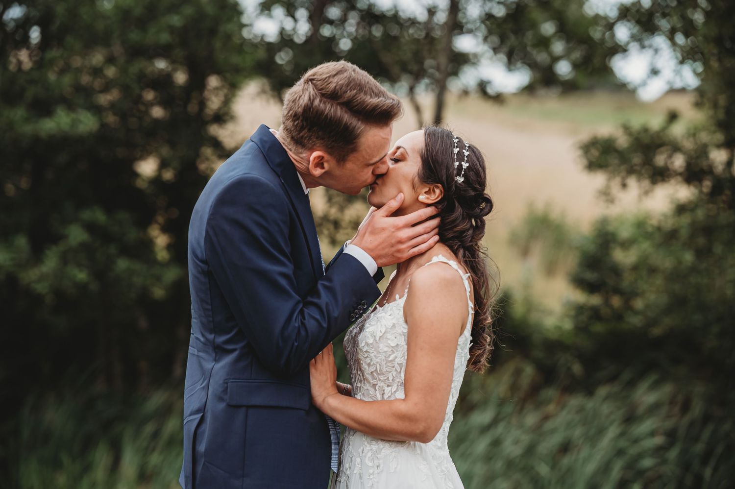 Romantic wedding couple sharing a tender kiss outdoors with natural greenery in the background.