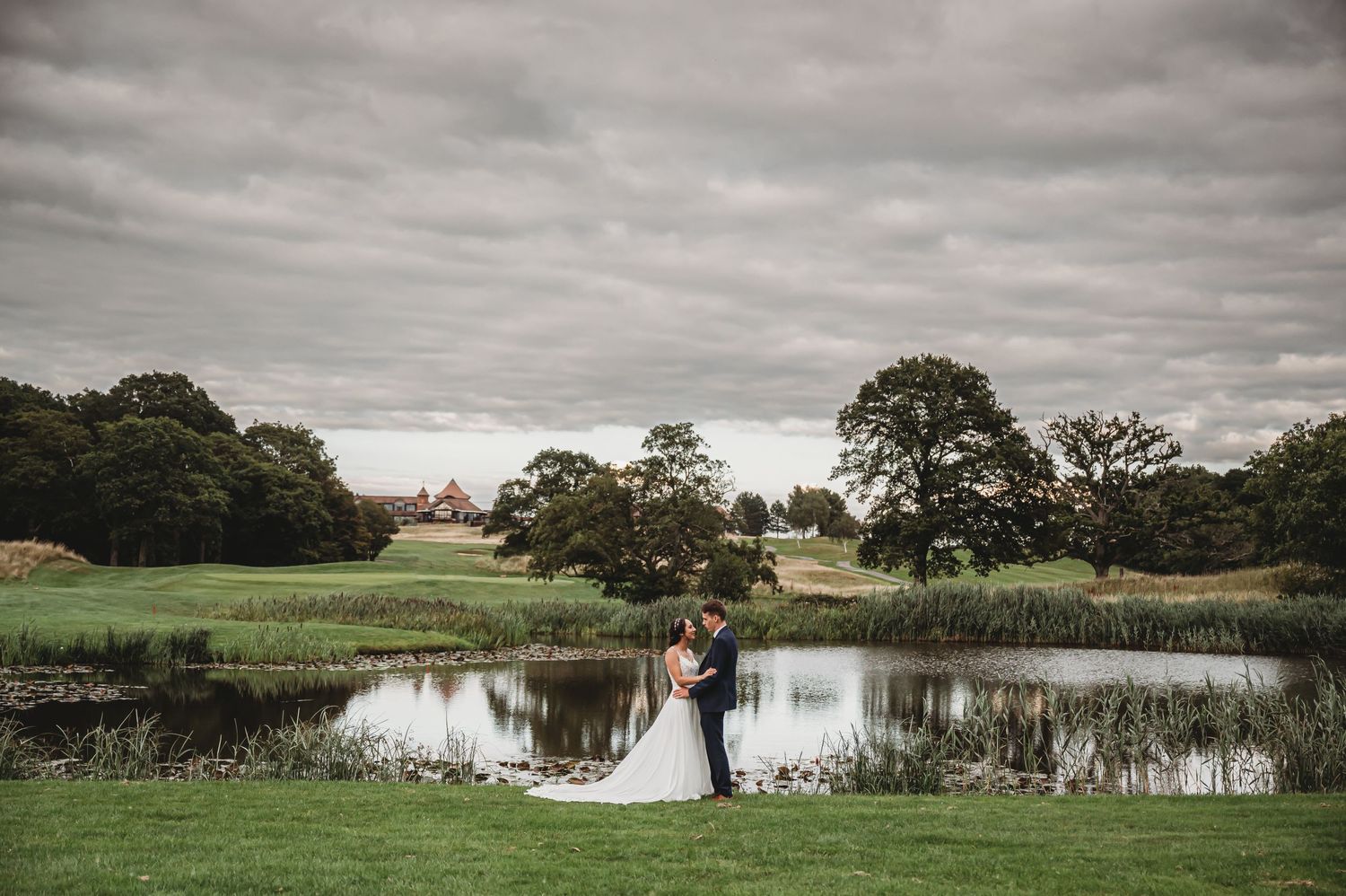 A couple shares a romantic moment by a serene pond surrounded by lush greenery under moody overcast skies.