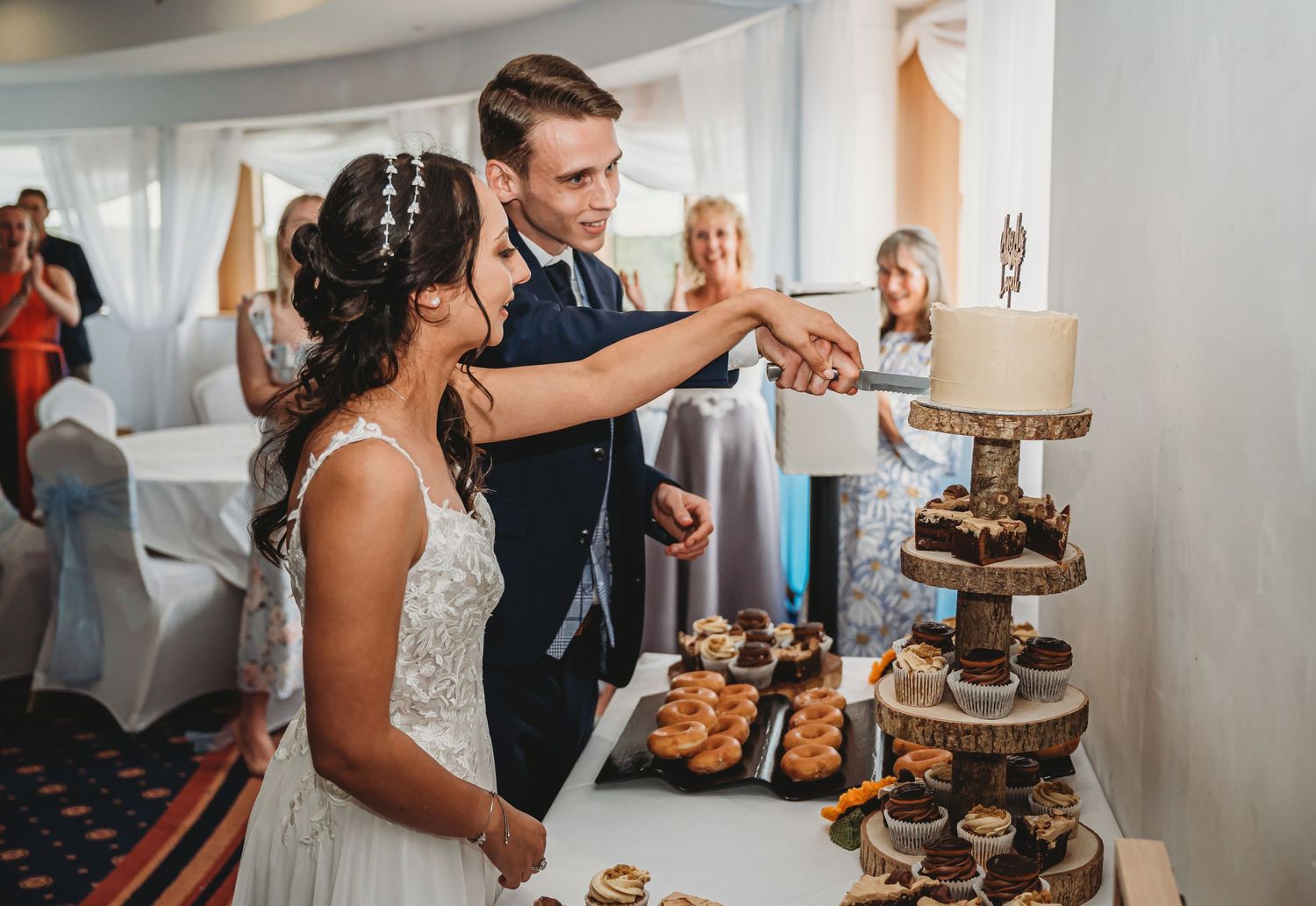A couple cuts their wedding cake together at their reception while guests look on in the background.