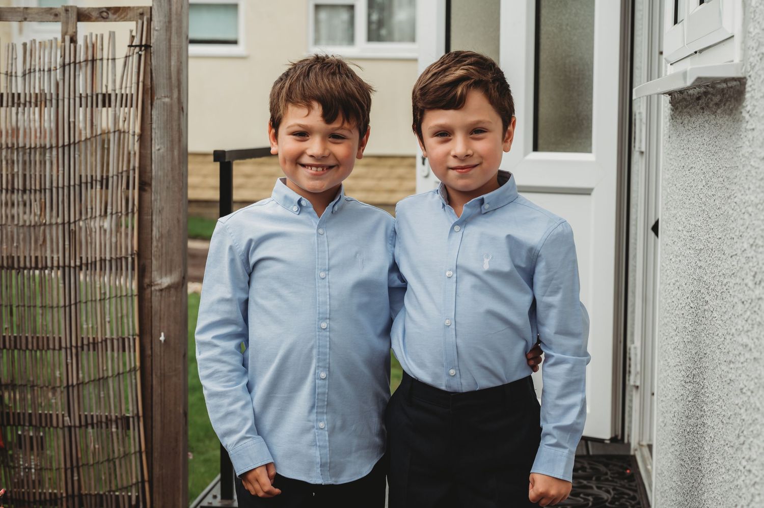 Two children wearing matching light blue button-down shirts and dark pants stand together outside a home.