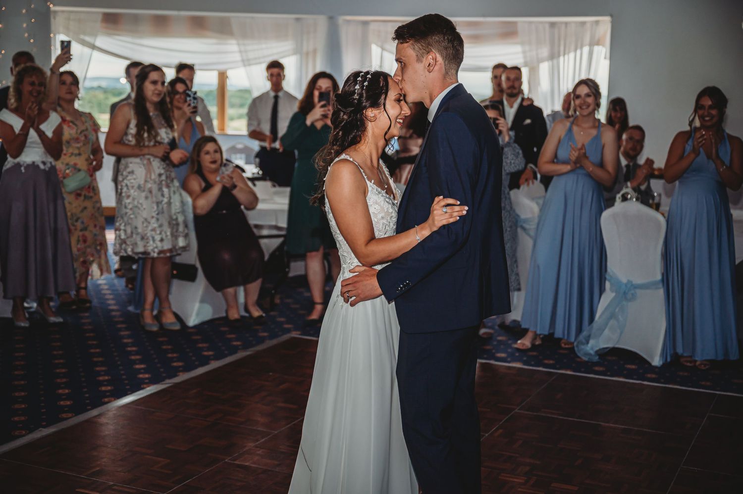 A couple shares their first dance at a wedding reception while guests in formal attire watch and applaud.