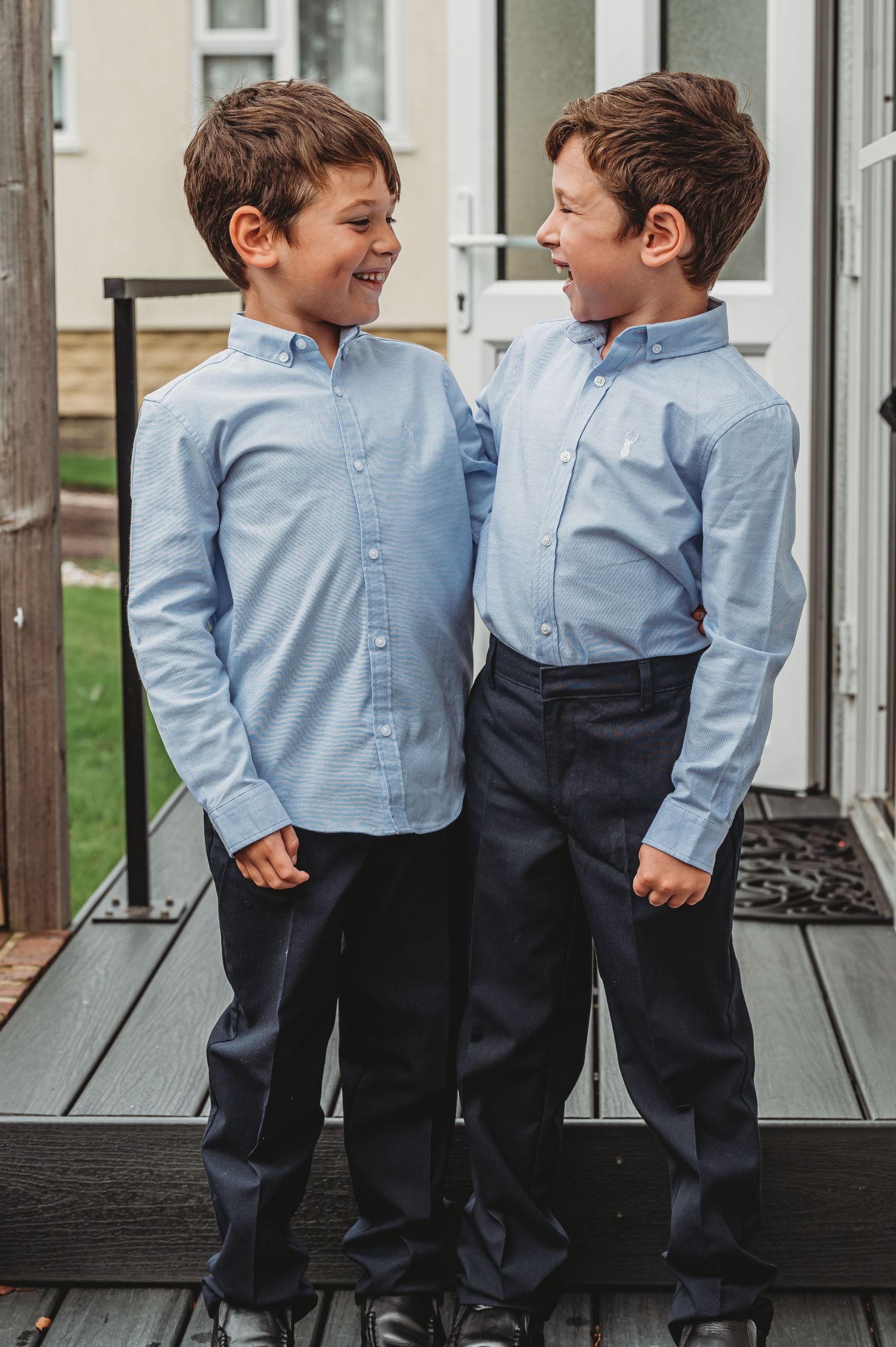 Two young children in matching light blue button-down shirts and navy pants stand together smiling on a wooden deck.