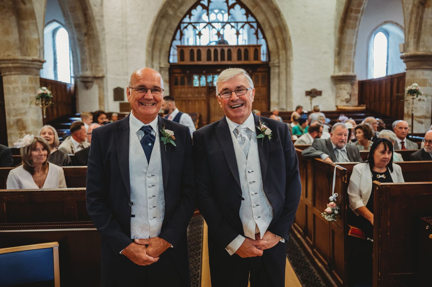 Two people wearing matching gray tuxedos stand together smiling at a church wedding ceremony.