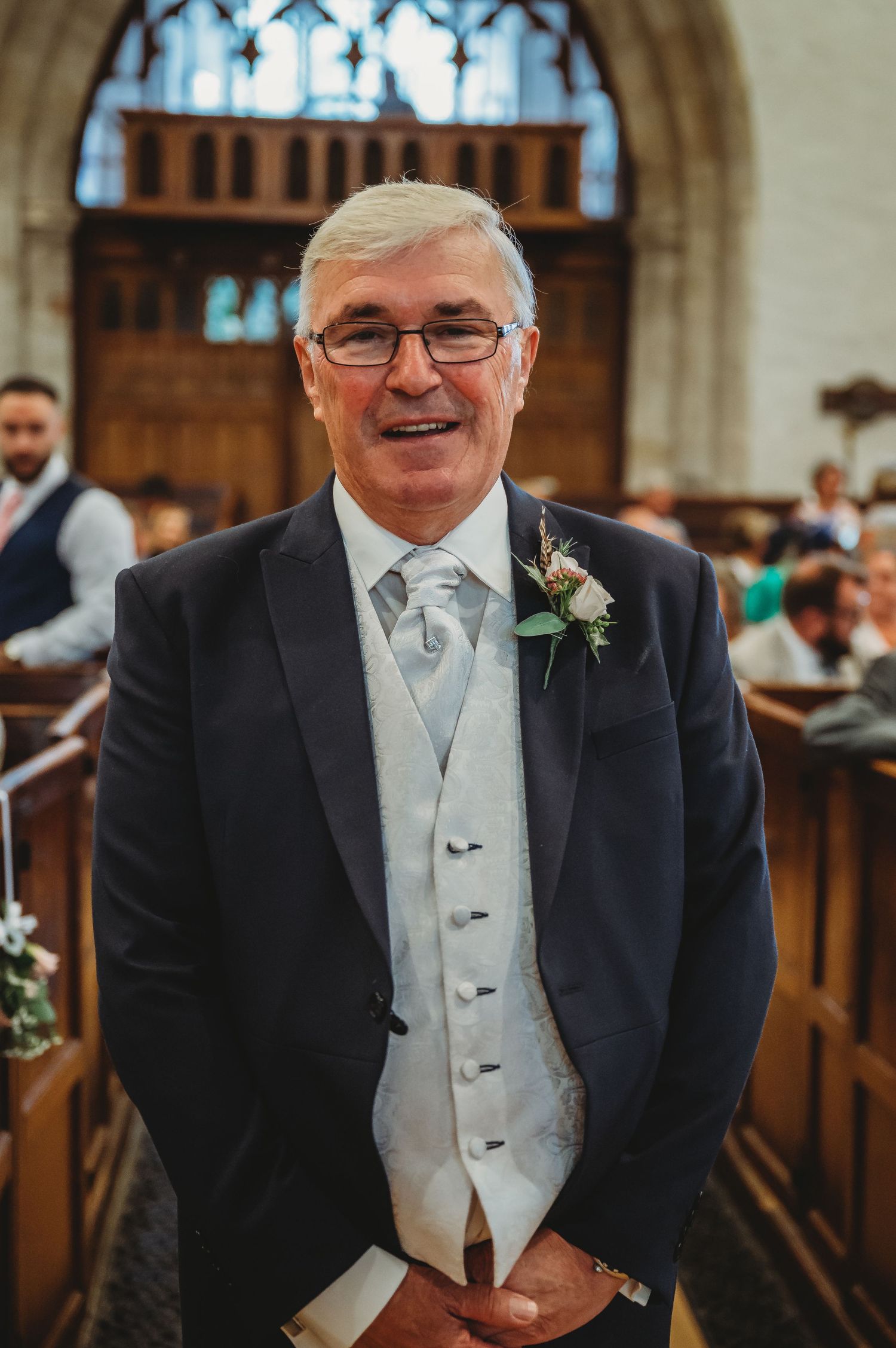 A person wearing formal wedding attire in a church with navy suit, light waistcoat and boutonniere.
