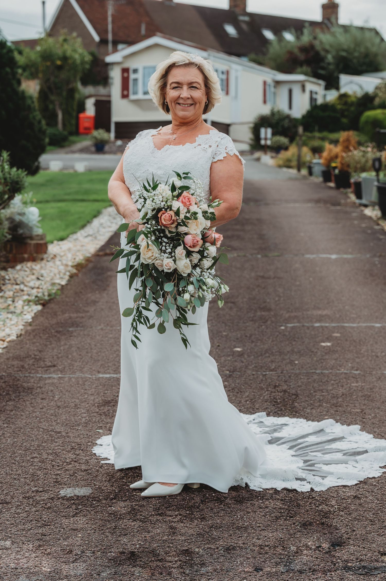 A person in an elegant white lace wedding gown holds a beautiful cascading bouquet of pink and white flowers on a path.