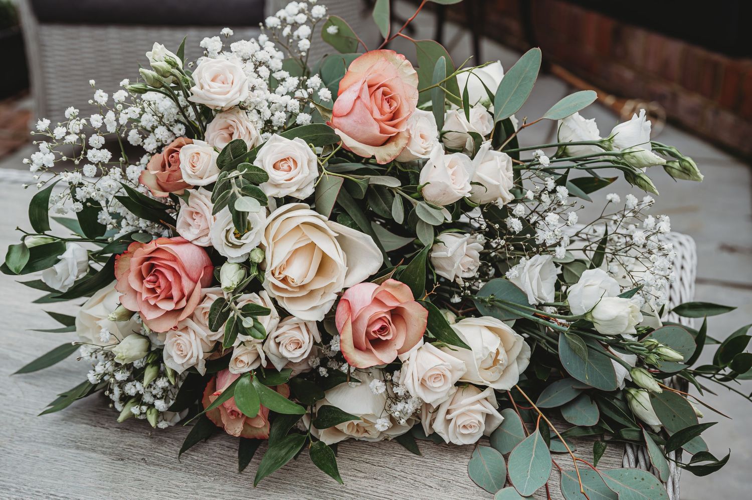 A romantic wedding bouquet with pink and cream roses, baby's breath, and eucalyptus leaves arranged on a gray surface.