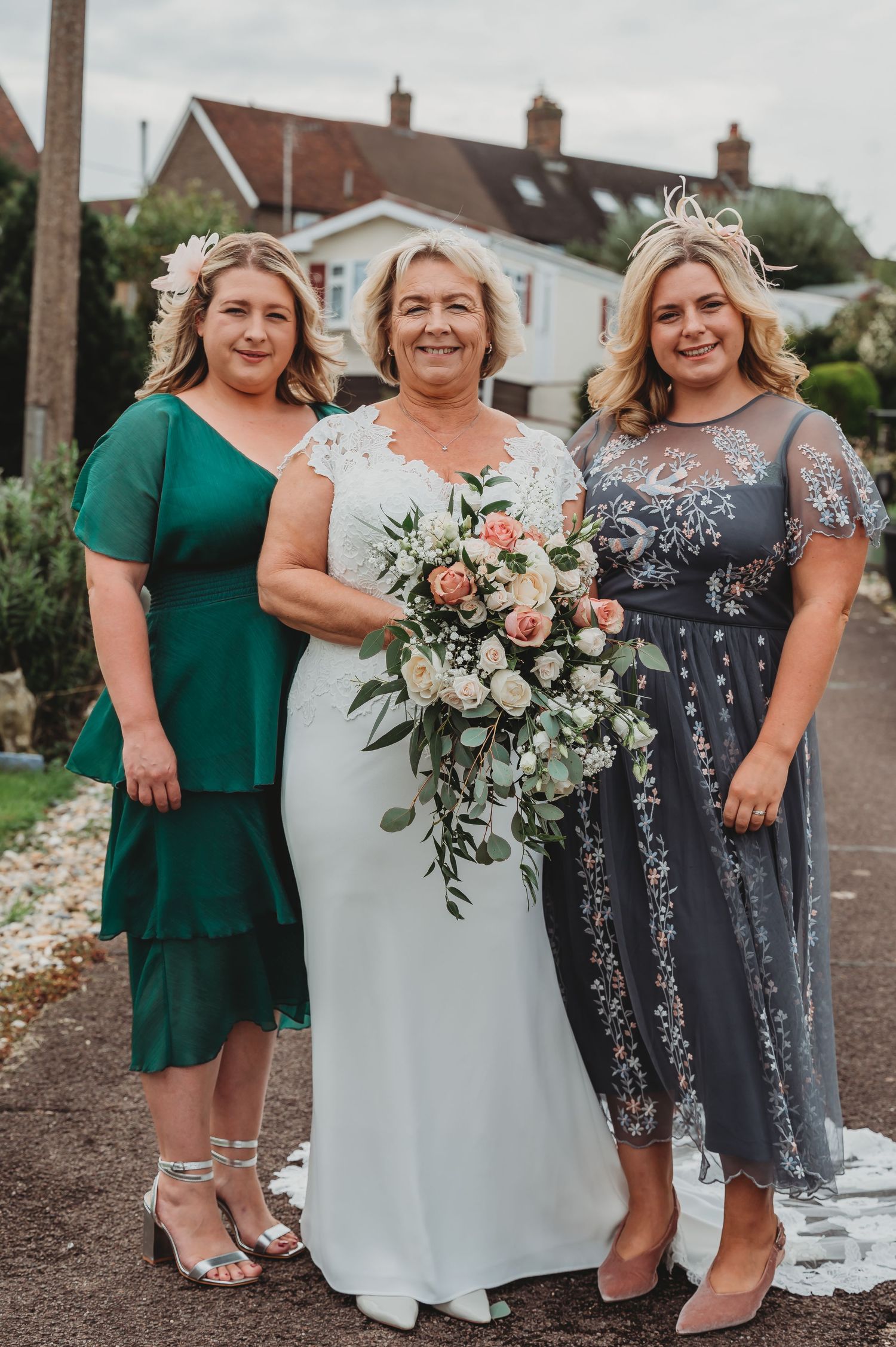 Three women in elegant dresses pose together at a wedding, with floral bouquet and suburban homes in background.