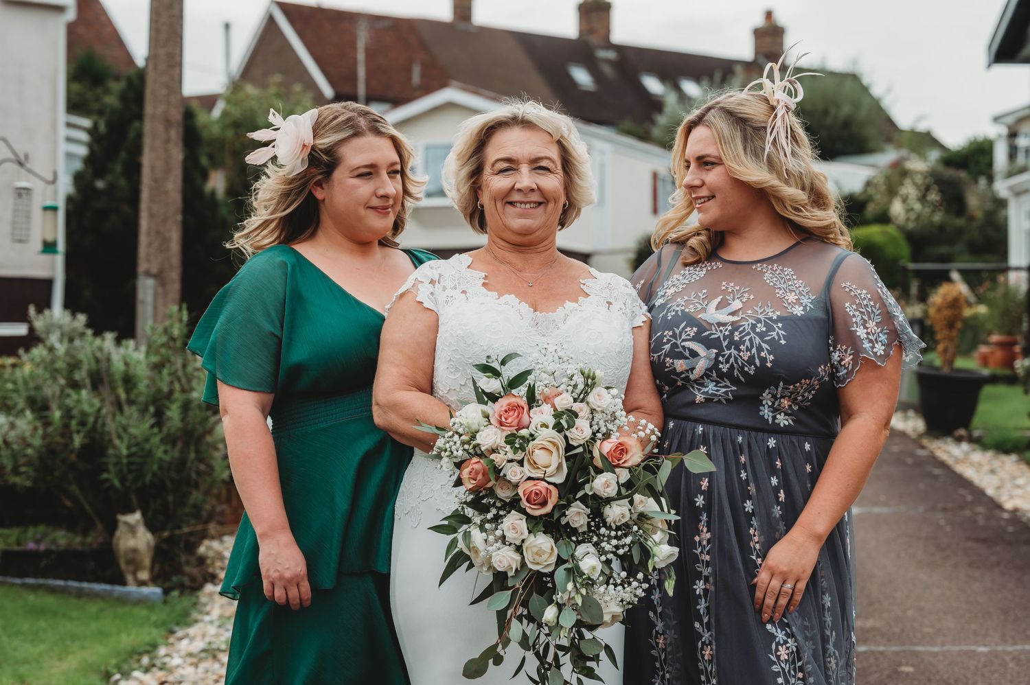 Three people wearing formal dresses smile together during a wedding ceremony, with one holding a floral bouquet.