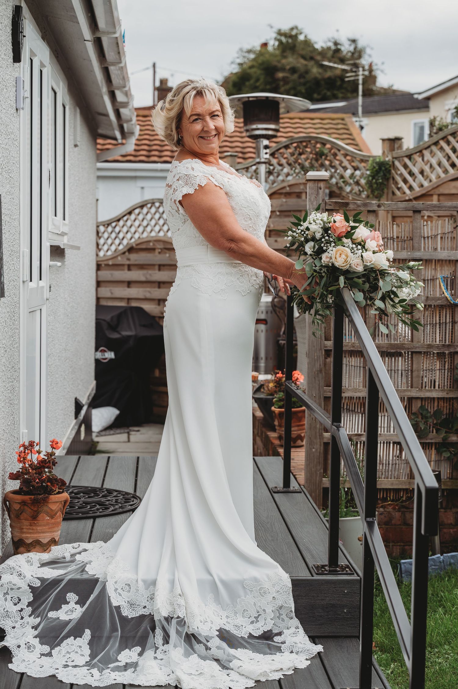 Beautiful bridal portrait on a garden deck wearing an elegant white lace gown with a flowing train and holding a floral bouquet.