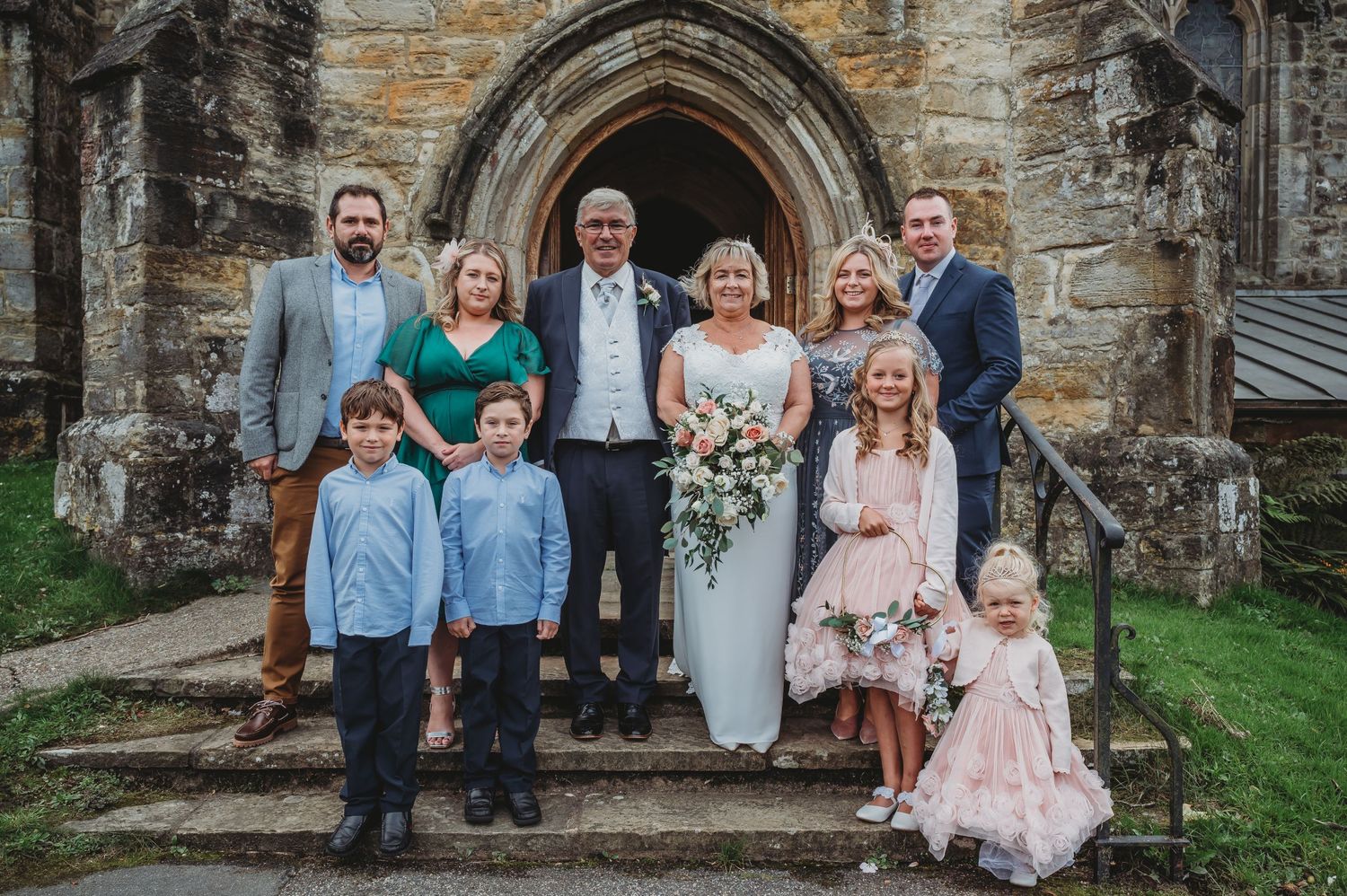 A wedding party poses on stone church steps in front of an arched Gothic doorway.